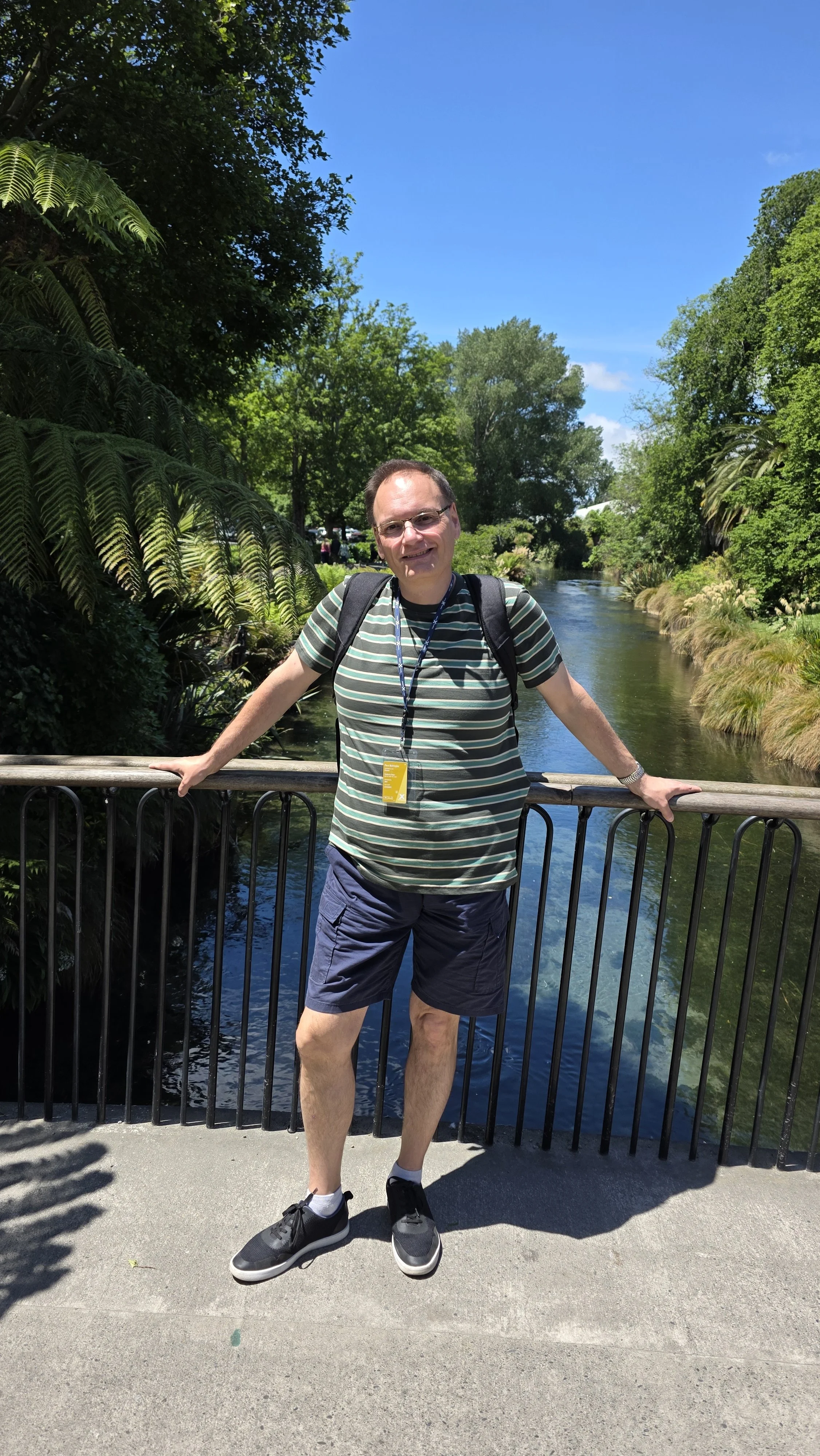 A man standing on a bridge over a river, with trees and greenery in the background, wearing a striped shirt, shorts, and sneakers, smiling for the camera.