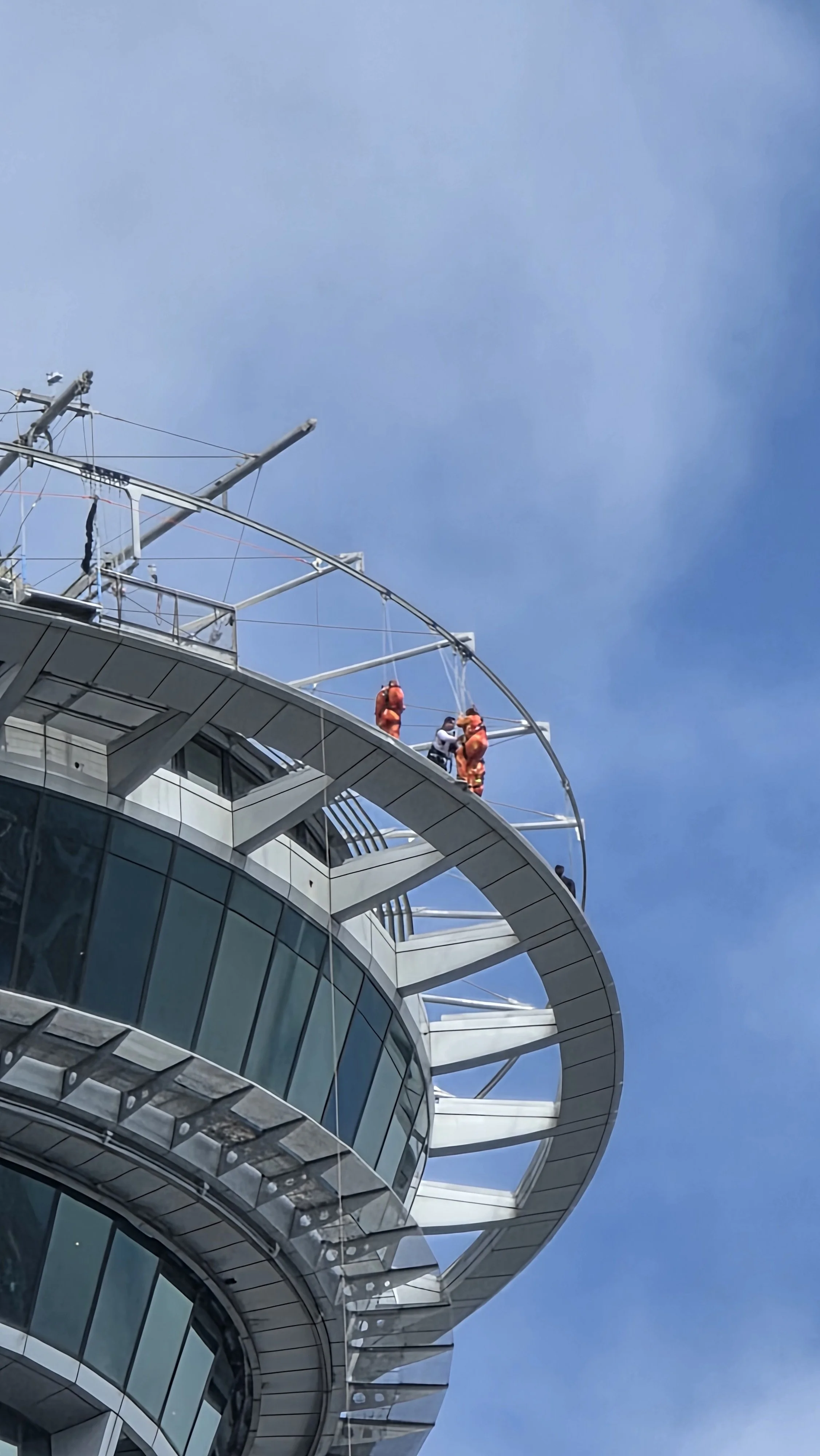 Construction workers wearing orange safety suits working on the top of a modern glass building with a circular observation deck against a blue sky.