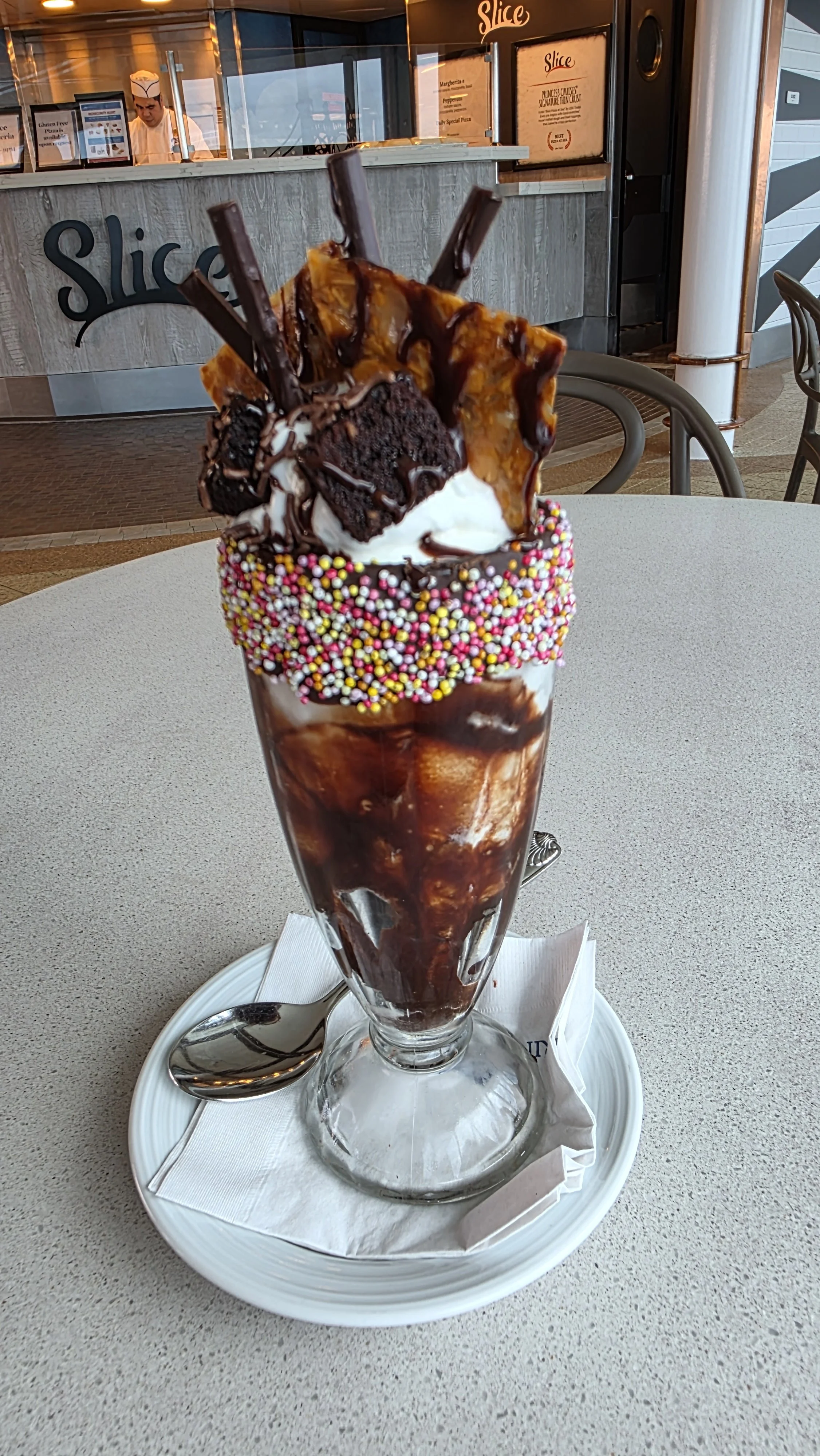 A tall ice cream sundae with whipped cream, chocolate syrup, rainbow sprinkles, chocolate wafer rolls, and a brownie piece, served in a tall glass.
