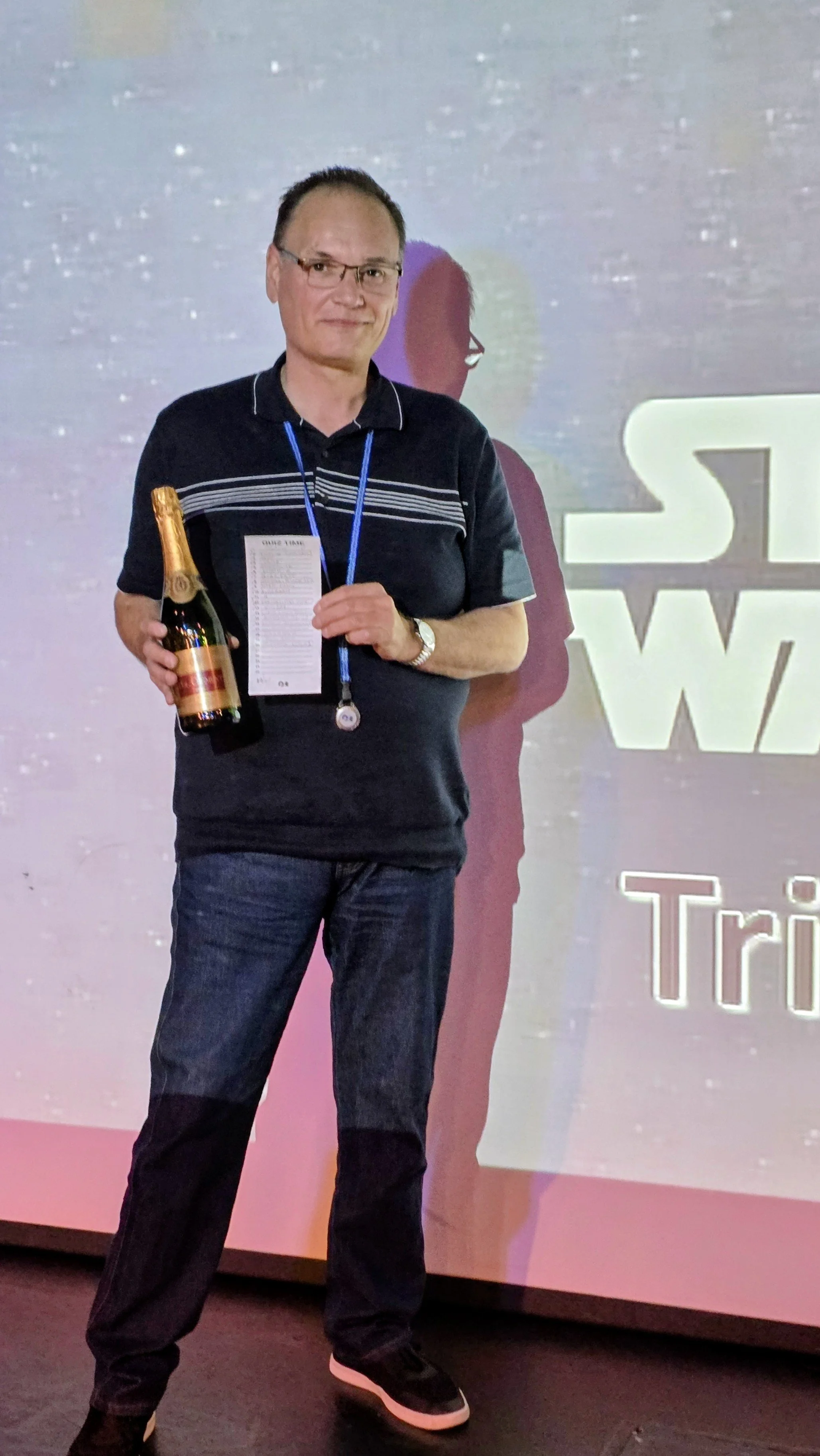 A man holding a bottle of champagne at an event with a large screen behind him displaying the words 'Star Wars' and 'Tri'. He is wearing glasses, a dark polo shirt with stripes, jeans, and sneakers, and has a medal around his neck.