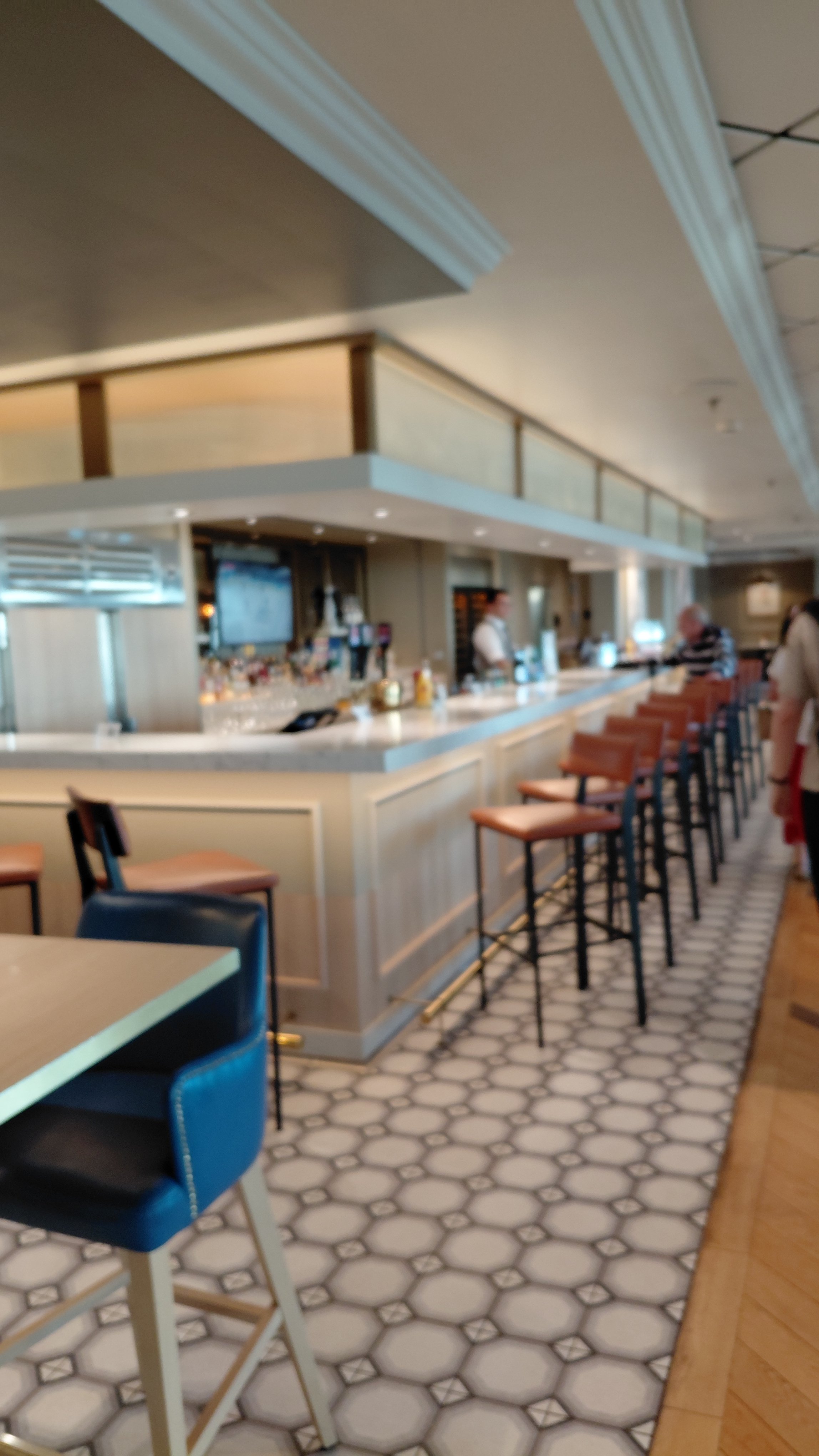 Inside a bar with a white marble countertop, bar stools, and patrons waiting or ordering drinks. The floor has a hexagonal tile pattern and the ceiling features crown molding.