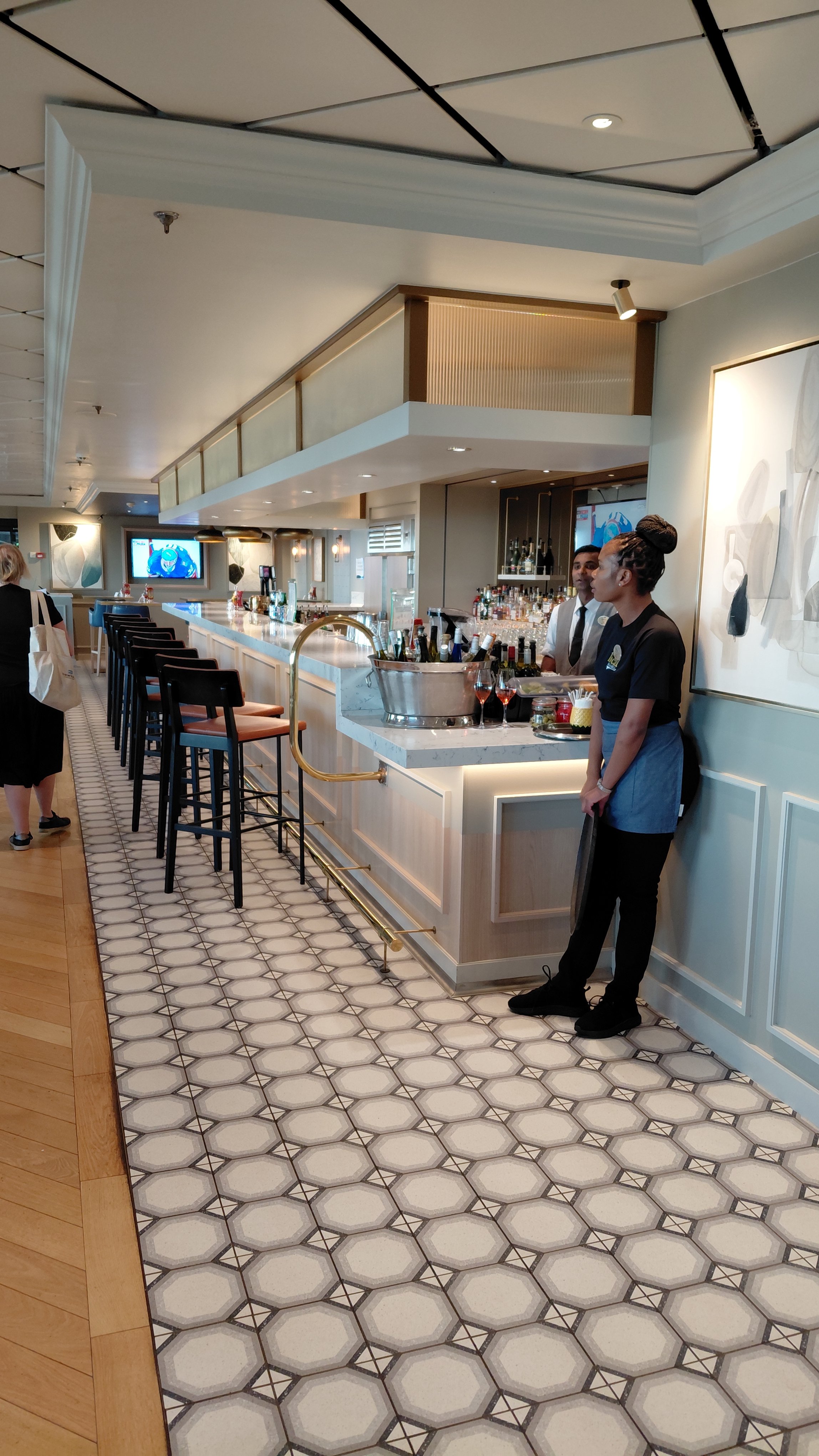 Bar area in a restaurant with black bar stools, liquor bottles behind the bar, and two staff members standing near the counter.