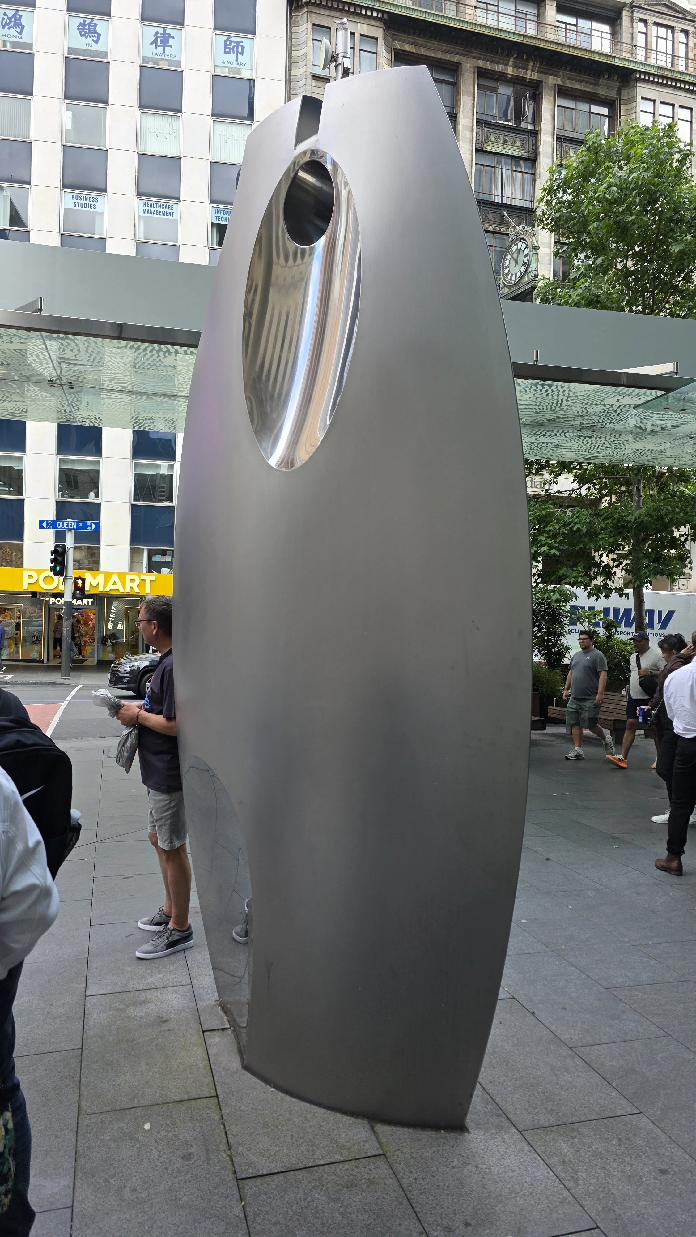 A large, modern, metallic sculpture with a circular cutout, standing on an urban sidewalk with people and city buildings in the background.