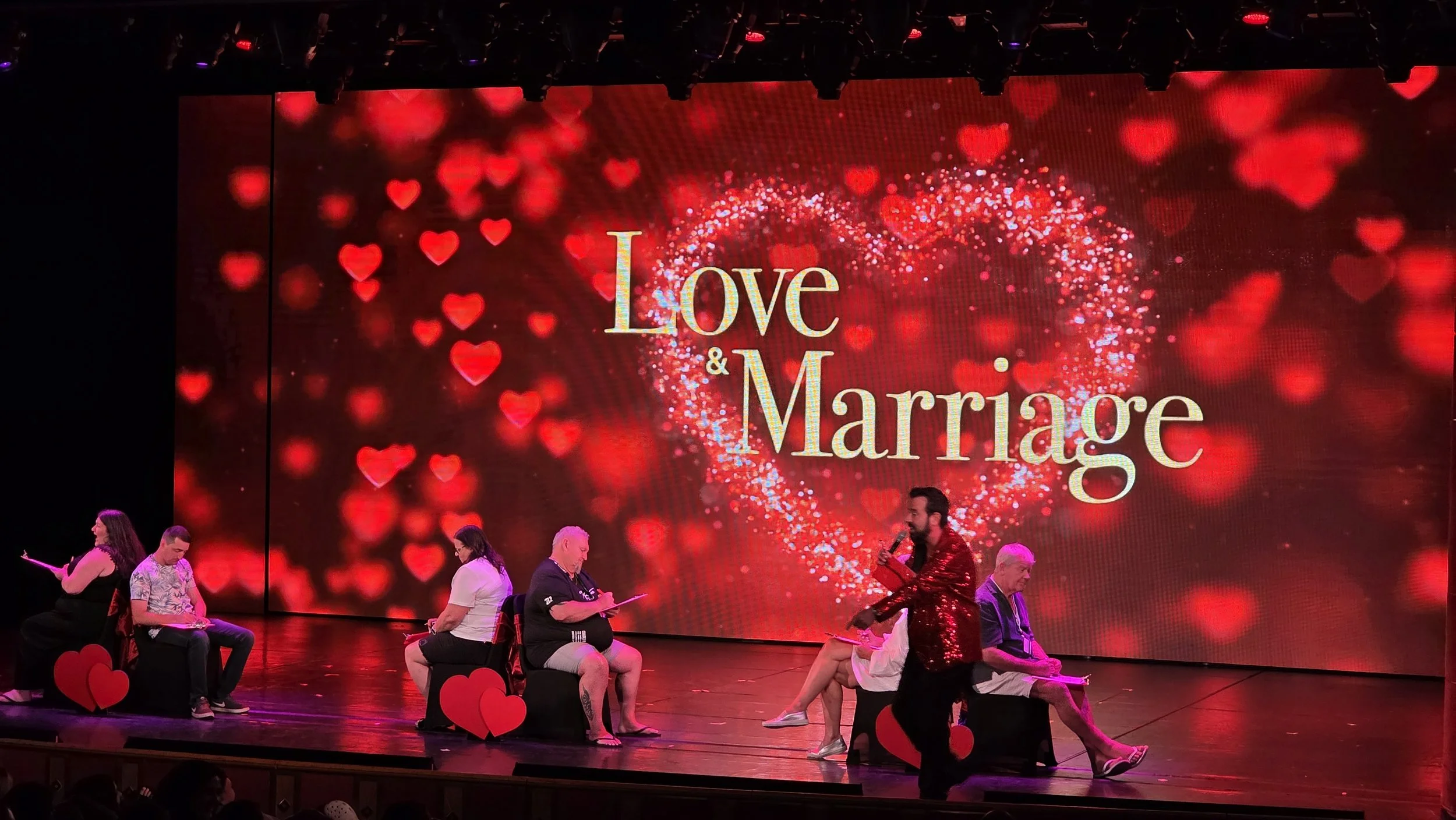 Stage with a large screen displaying a heart-shaped design and the words 'Love & Marriage' in champagne-colored text, surrounded by red hearts. Several people are sitting on stage chairs decorated with red hearts, and a man in a red sequined jacket i