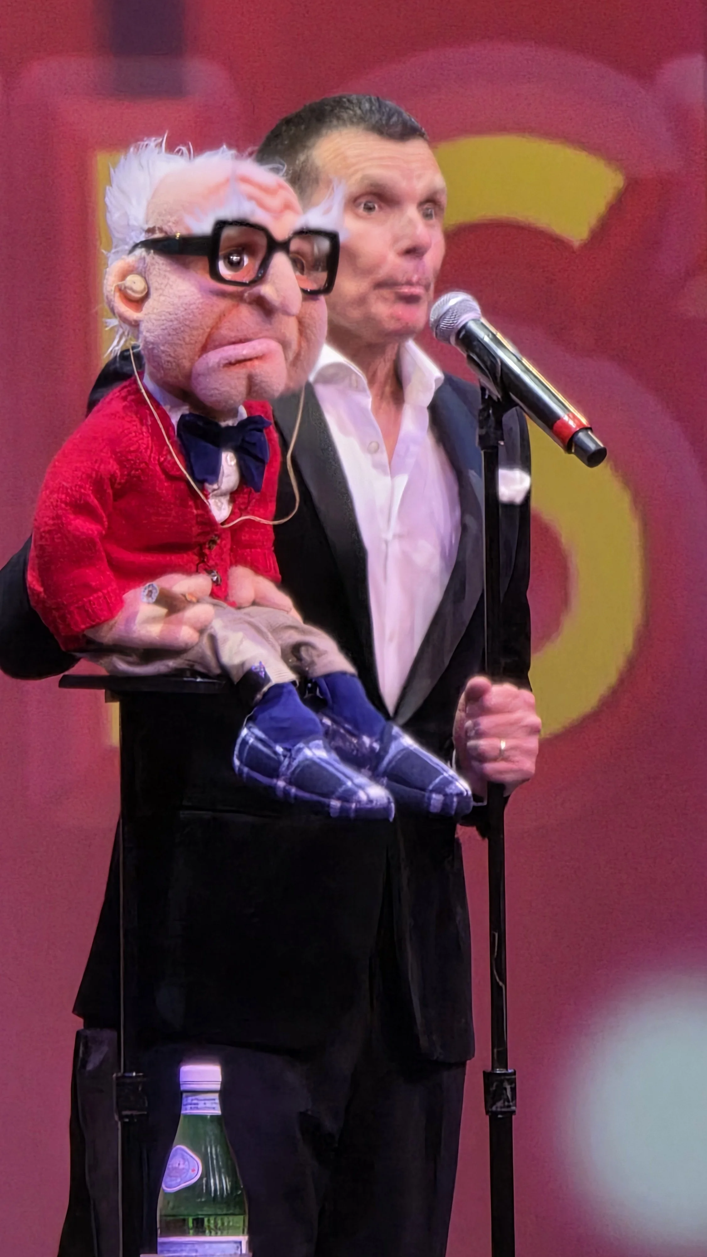 A man in a tuxedo holding a hand puppet with glasses, white hair, and a red sweater, standing behind a microphone on a pink and yellow background.