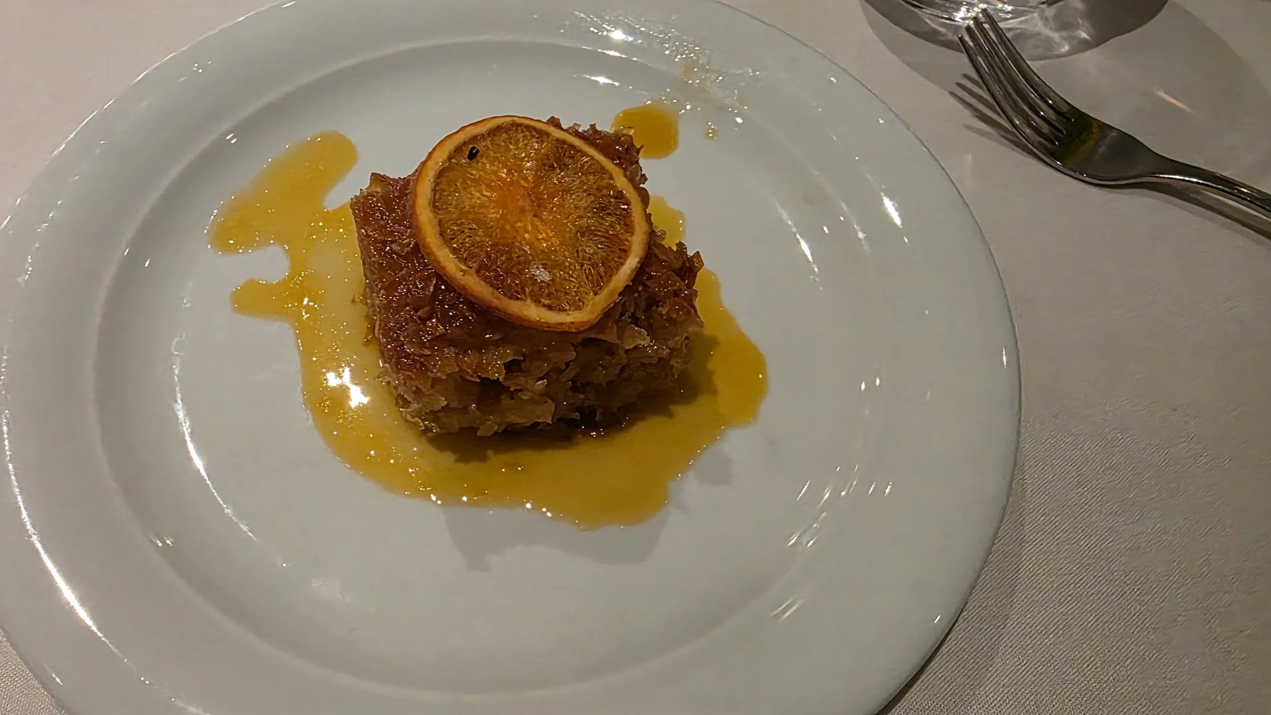 A plated dessert with a square of apple crumble topped with a dried orange slice and drizzled with caramel sauce on a white plate