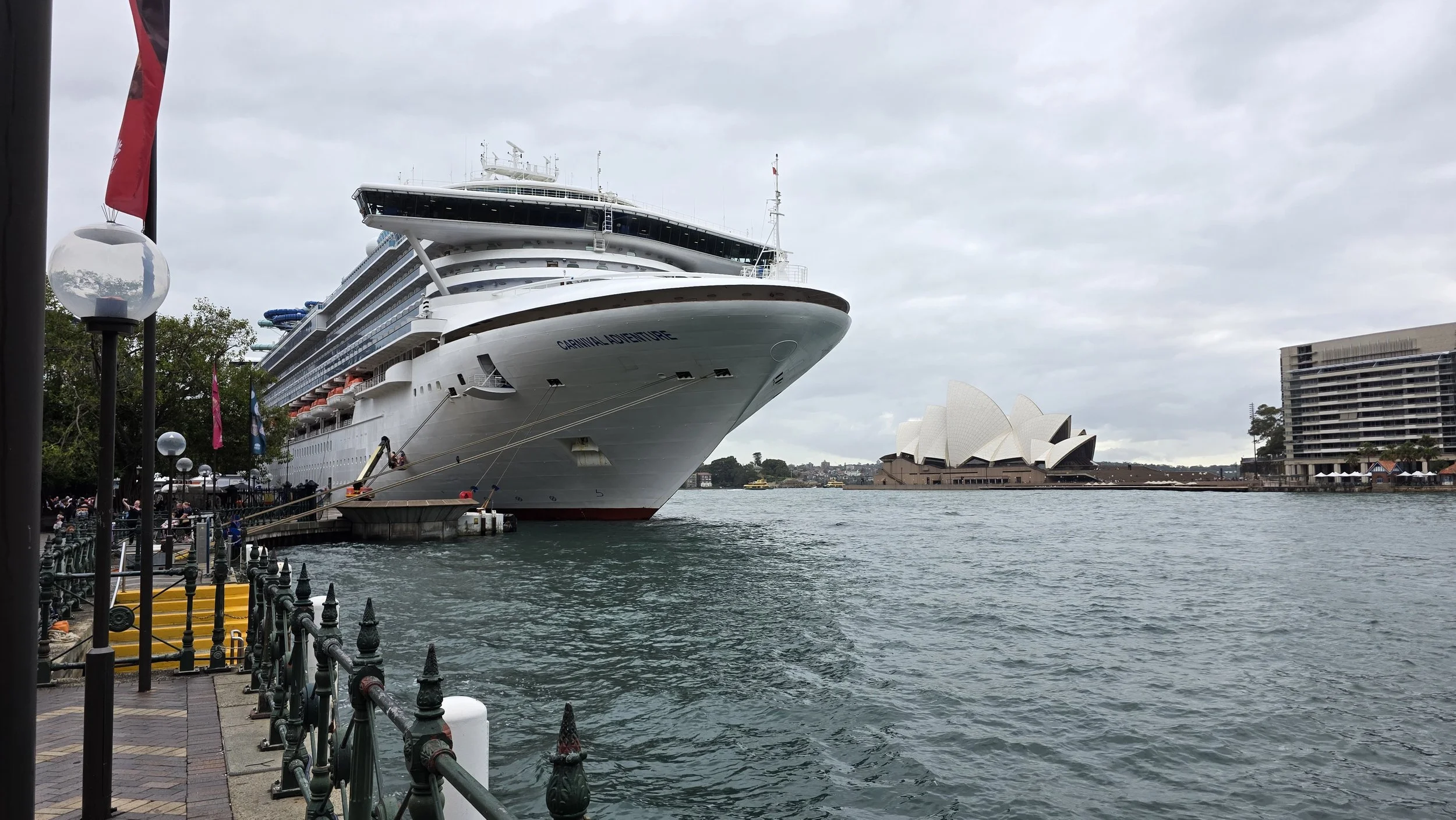 A large white cruise ship docked at the harbor with the Sydney Opera House in the background on a cloudy day.