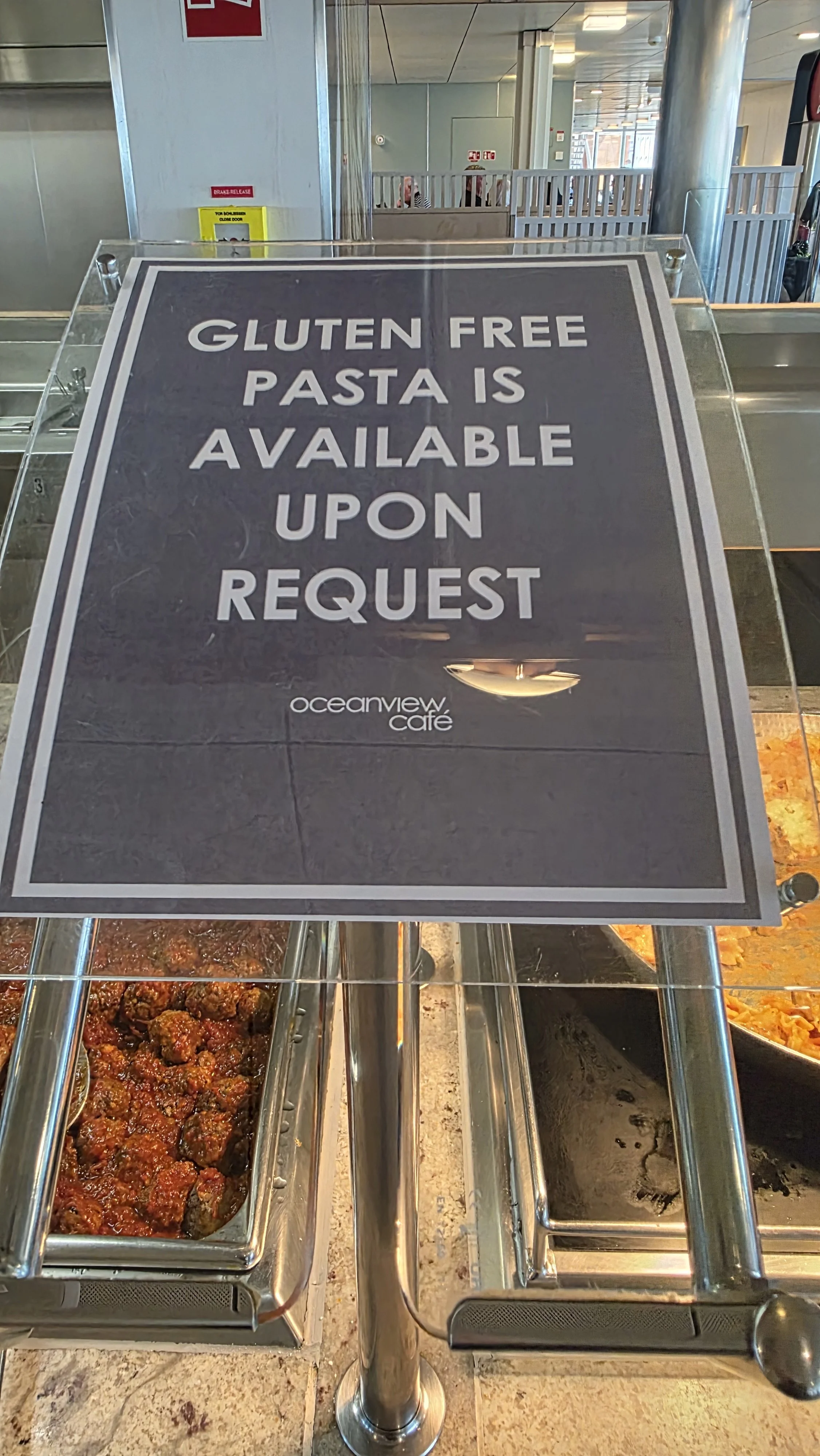 Sign at Oceanview Cafe advertising gluten free pasta available upon request, with trays of various food items underneath.