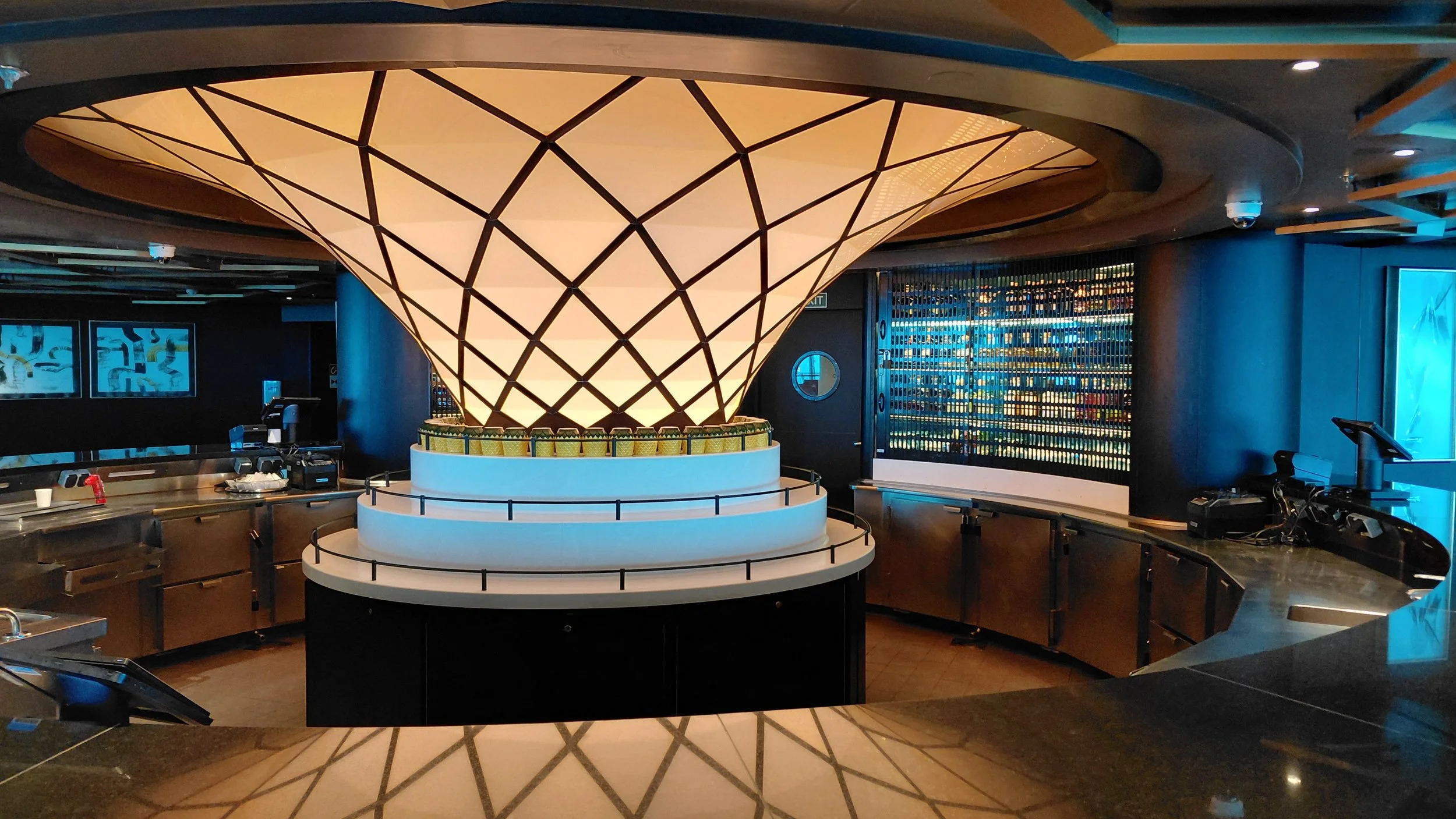 Interior of a modern bar or restaurant with a large illuminated geometric cone-shaped light fixture as the centerpiece, surrounded by a counter and drink shelves.