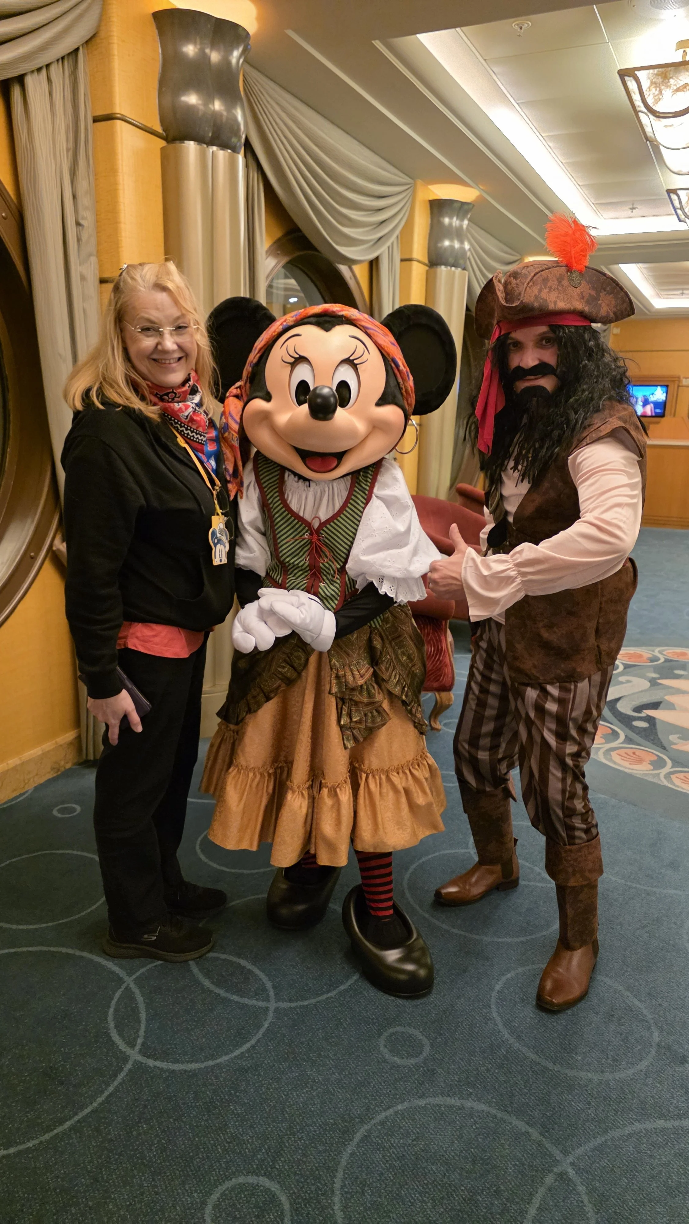 A woman standing with Mickey Mouse dressed as a pirate and another person dressed as a pirate in a carpeted indoor area with yellow walls, curtains, and decorative columns.
