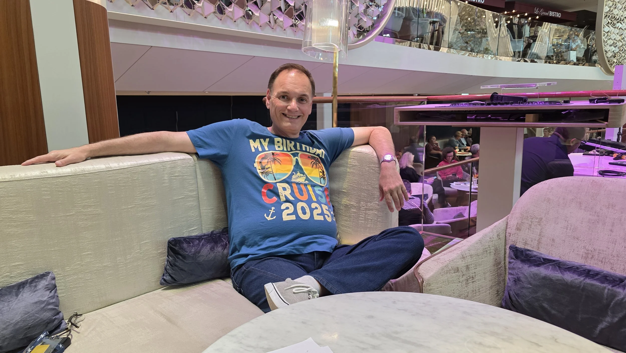 A man with short hair, wearing a blue T-shirt with a cruise graphic and the text "My Birthday Cruise 2023," sits on a cream-colored couch in a busy, brightly lit indoor space, smiling at the camera with his arm resting on the back of the couch.