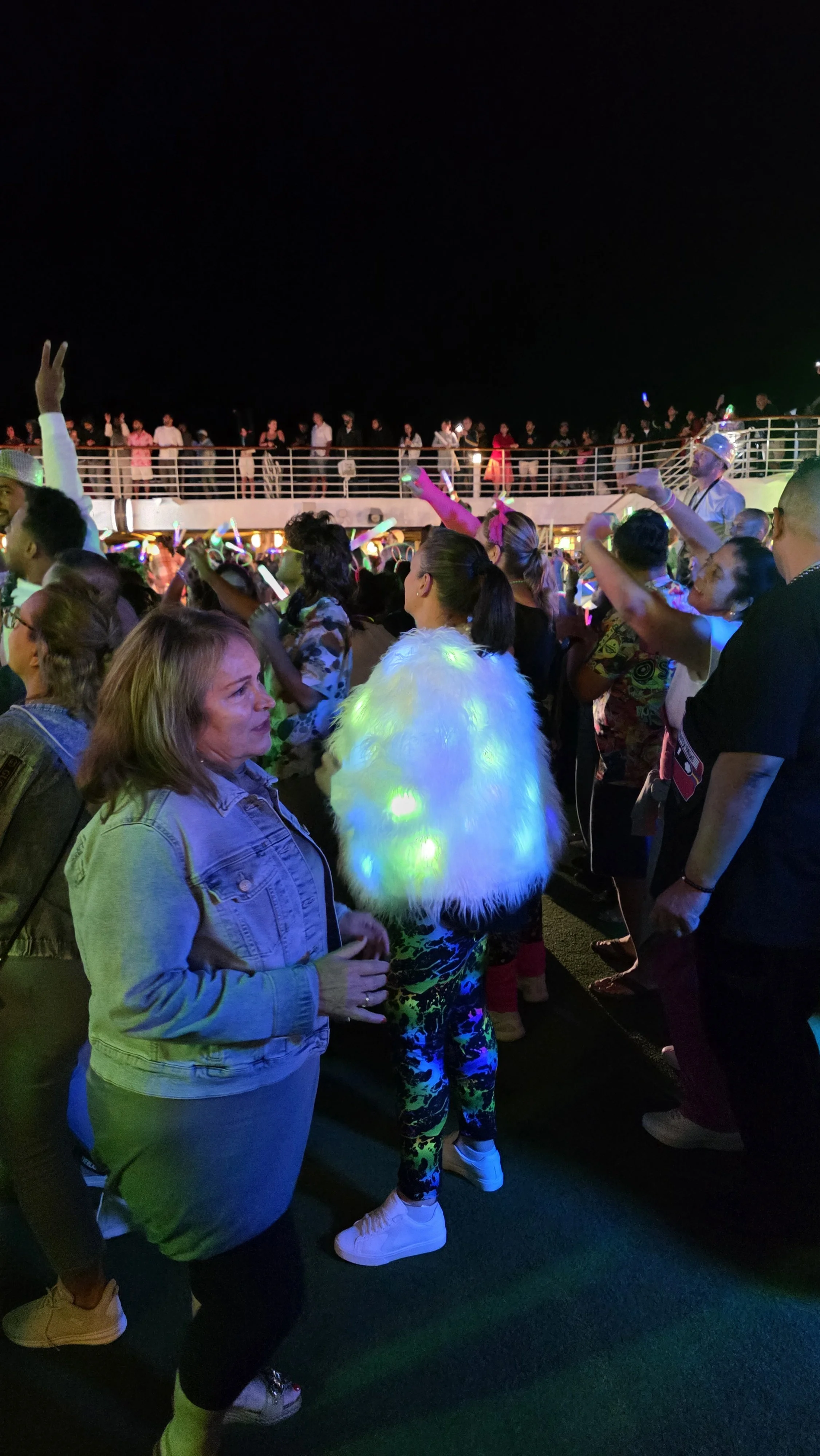 People dancing and socializing on a cruise ship at night, with a balcony and more people in the background.