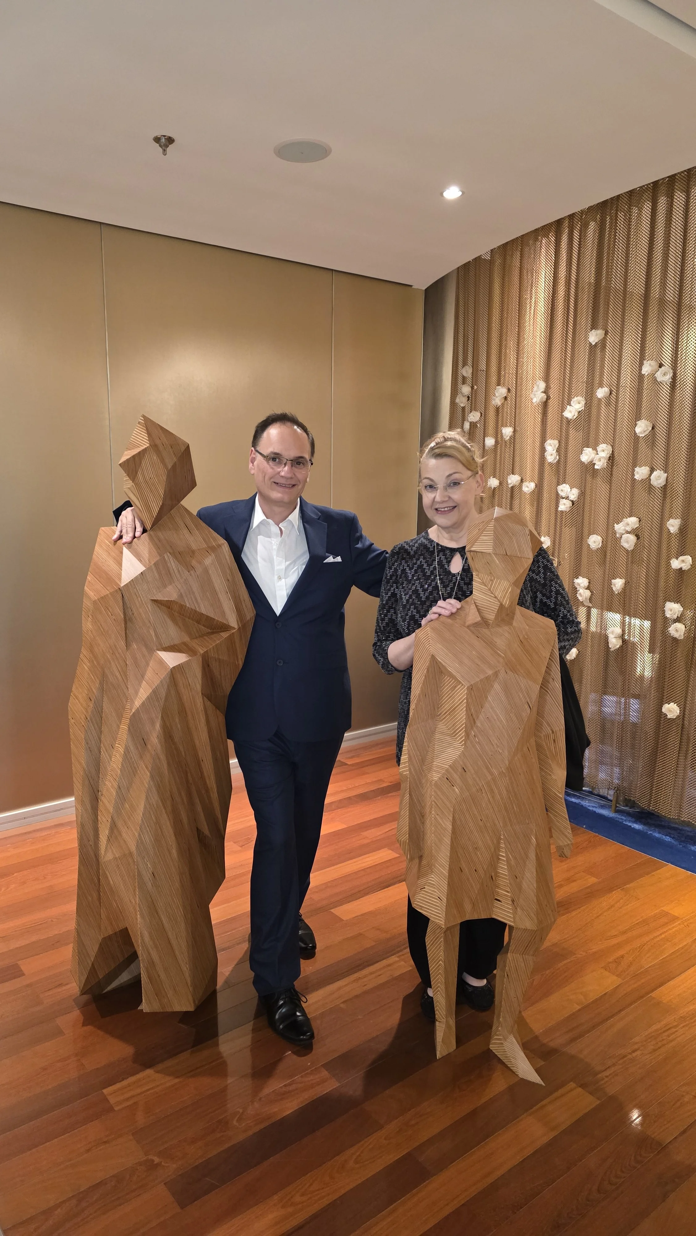 A man and a woman standing indoors, each with a wooden geometric sculpture resembling a human figure. The man is wearing a navy suit and glasses, and the woman is wearing a black patterned dress with glasses. They are smiling and posing for the photo