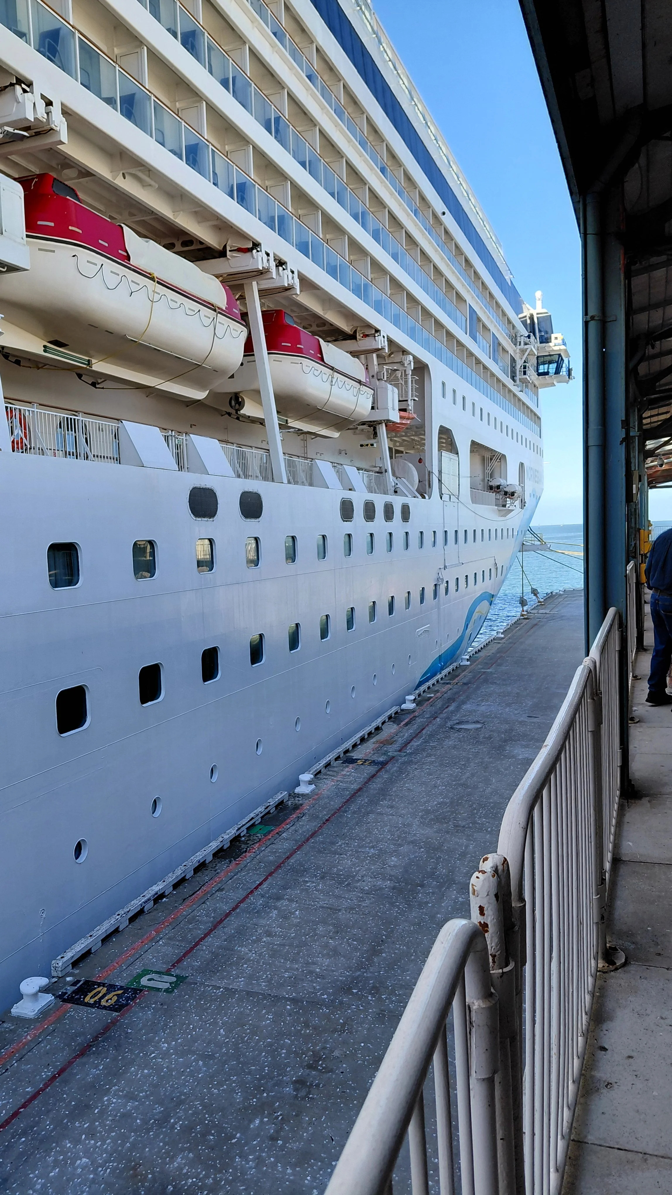 A large white cruise ship docked at a port, seen from the side with multiple decks and lifeboats attached.