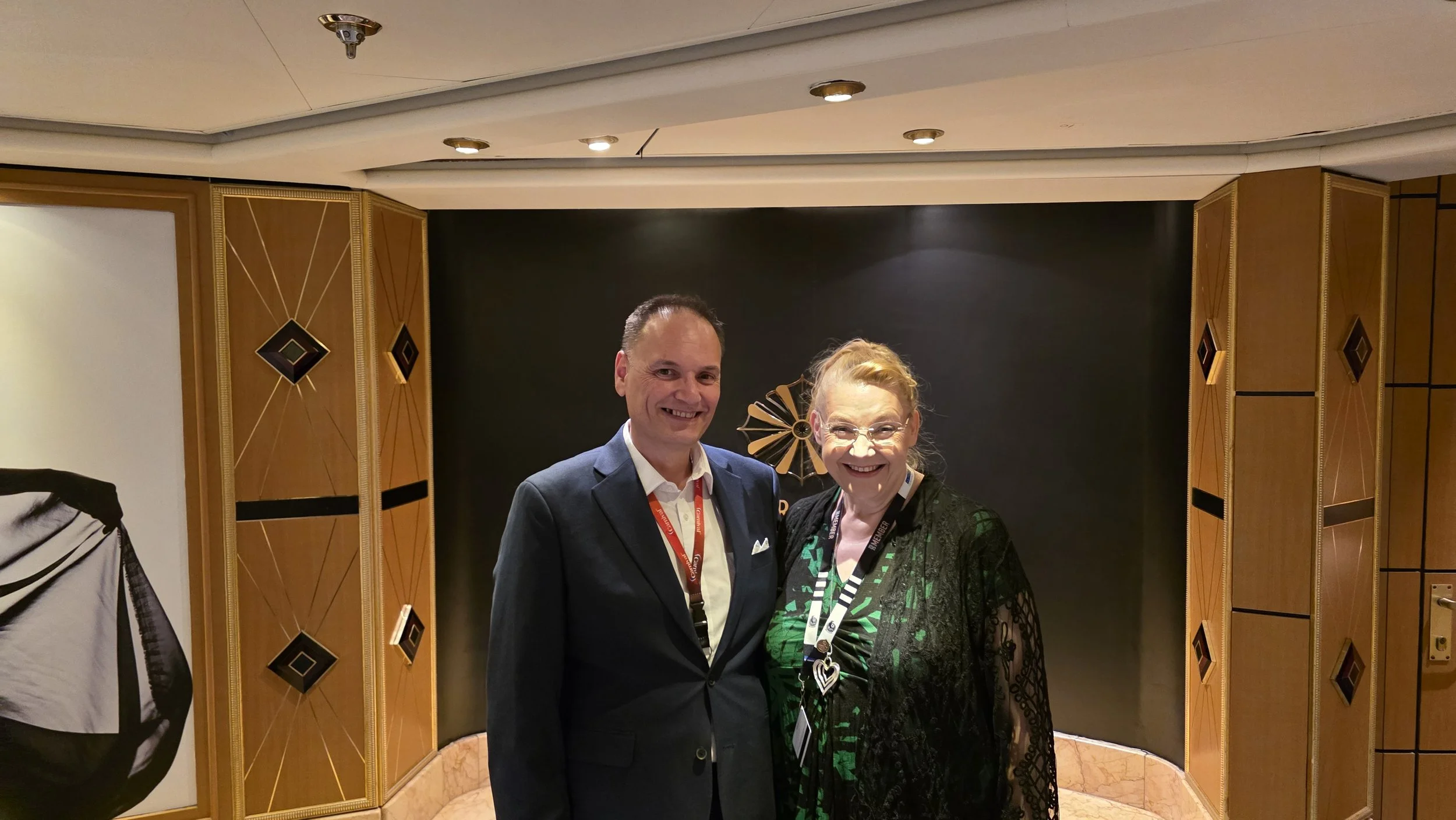 A man and woman standing side by side, smiling, in a hallway with decorative wooden panels and a black backdrop behind them.