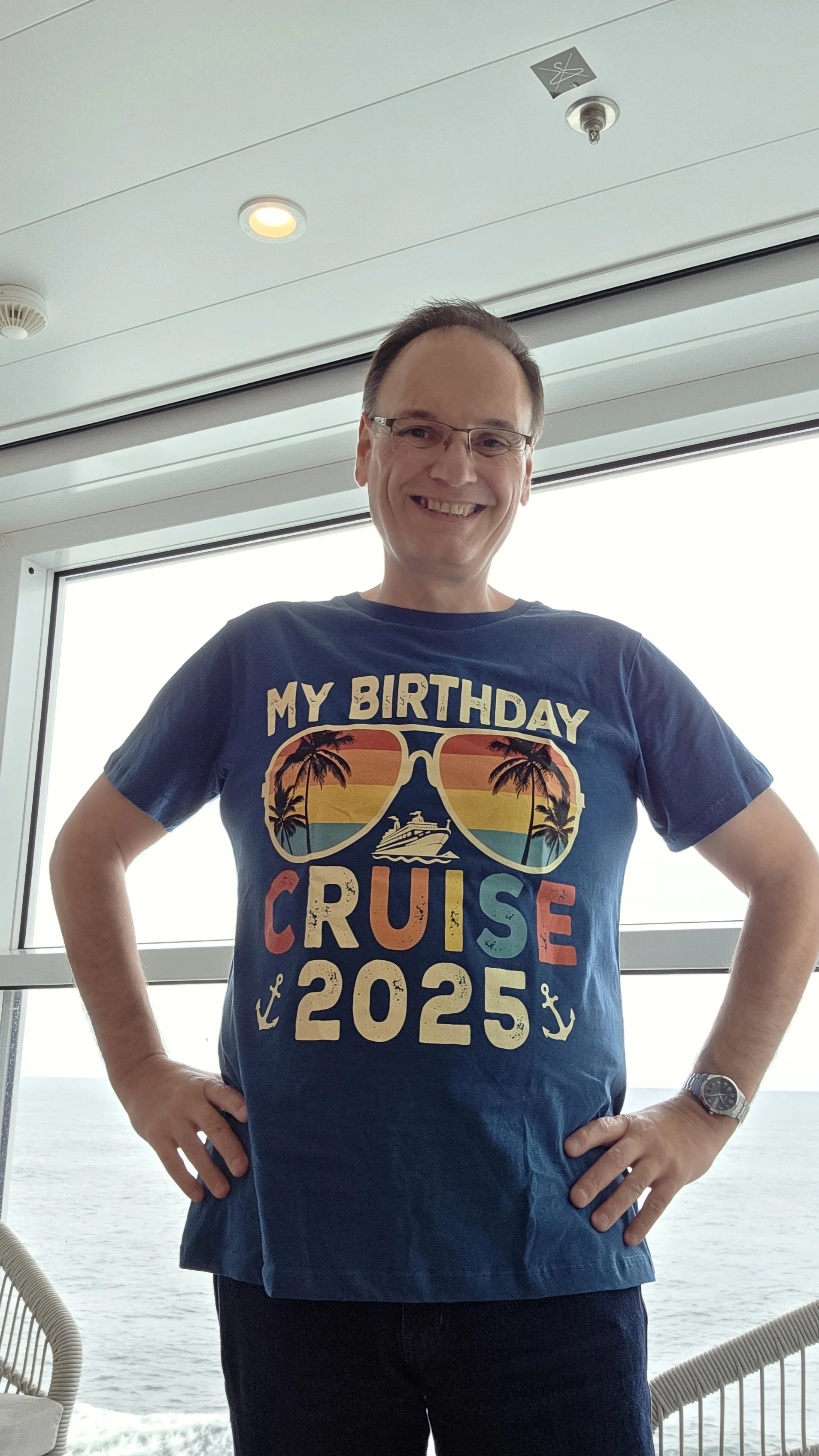 A man smiling while standing inside a cruise ship with a large window showing the ocean in the background. He's wearing a navy T-shirt that reads "My Birthday Cruise 2025" with palm trees, sunglasses, a cruise ship, and anchors on it.