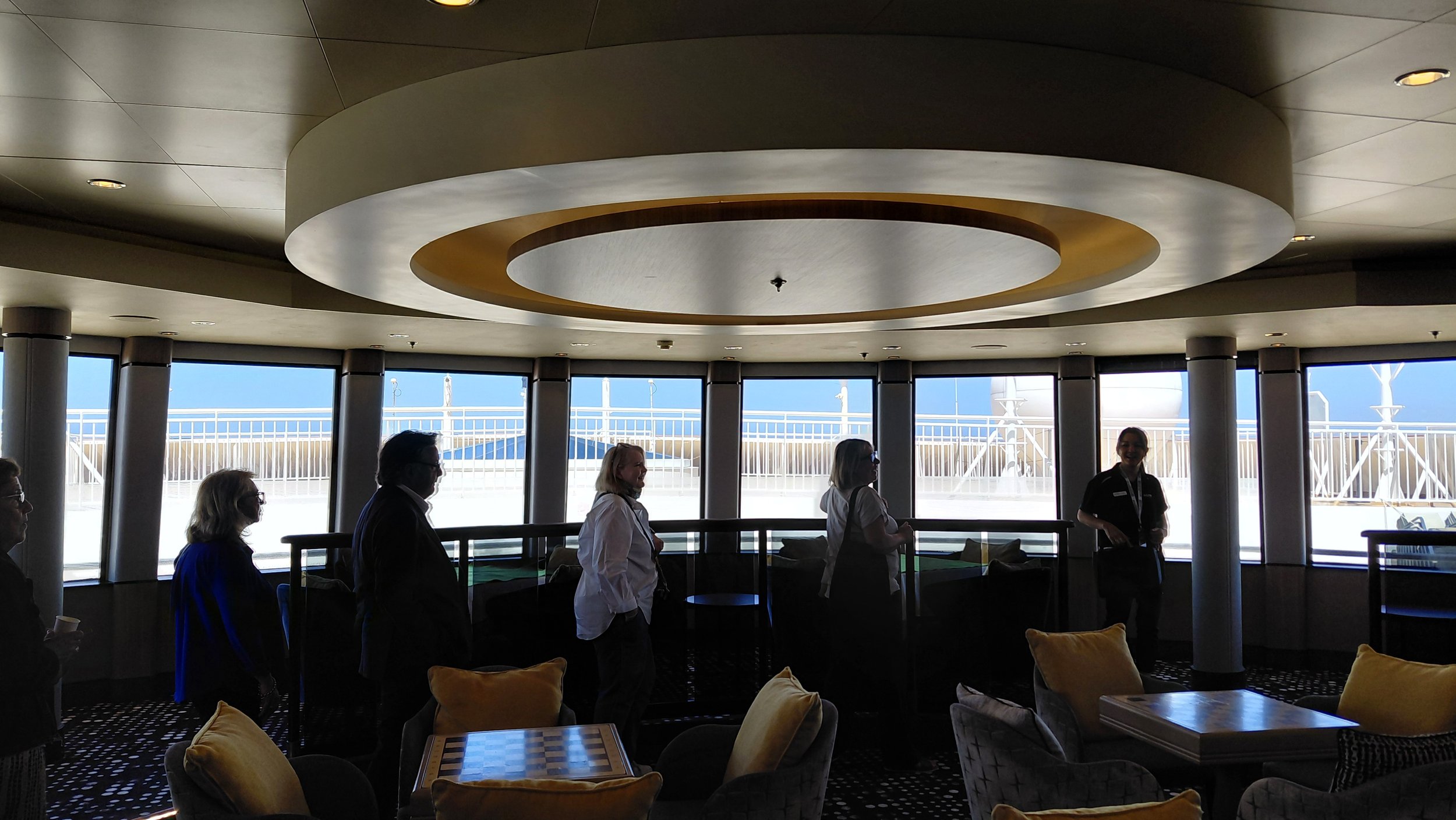 People standing inside a cruise ship's indoor lounge area near large windows with a view of the outdoor deck and sky, with seating and tables in the foreground.