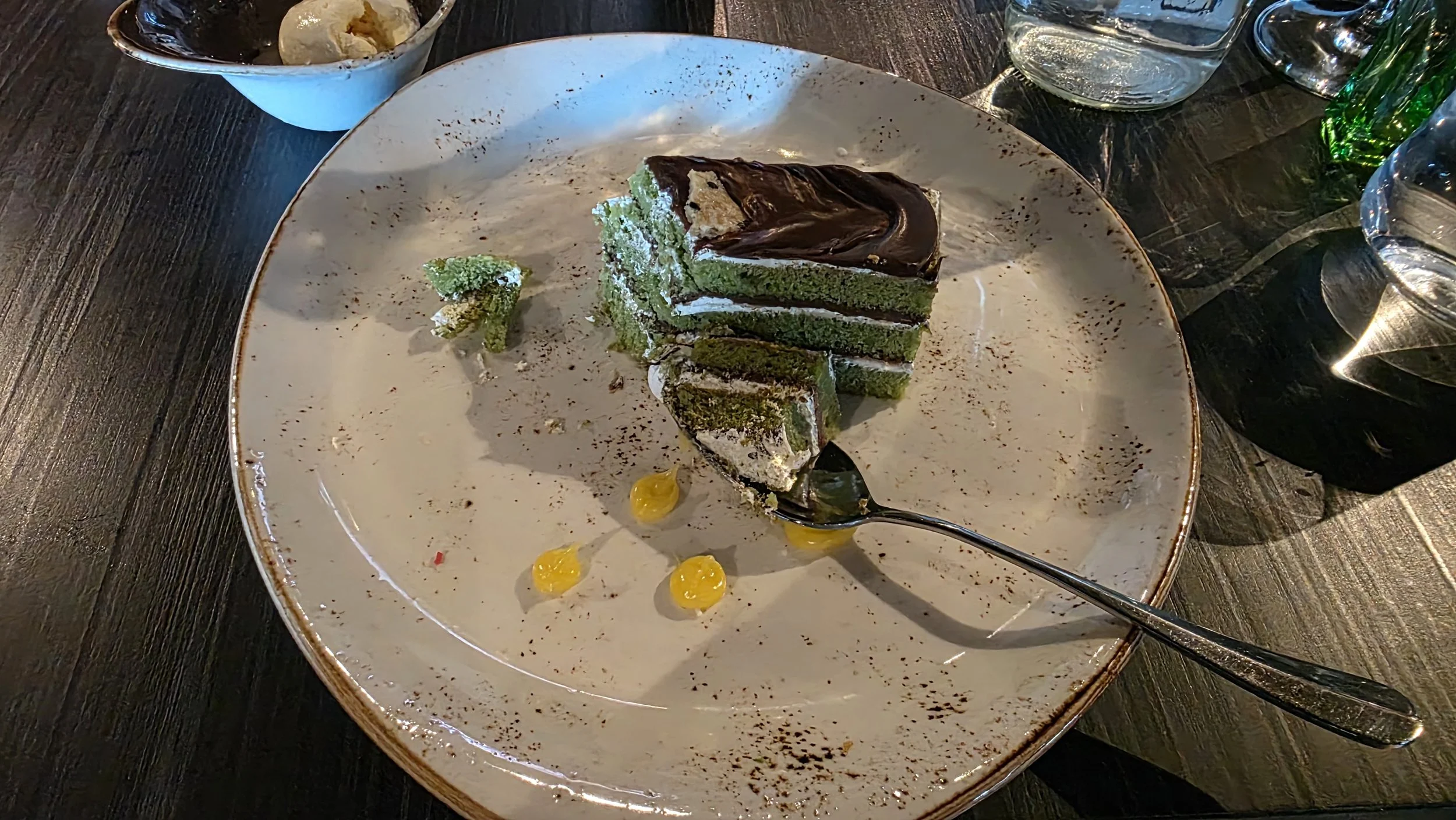 A slice of green layered cake with chocolate glaze on top on a white plate, accompanied by green jelly candies, a small piece of broccoli, and a spoon. There are glasses of water on the dark wooden table.