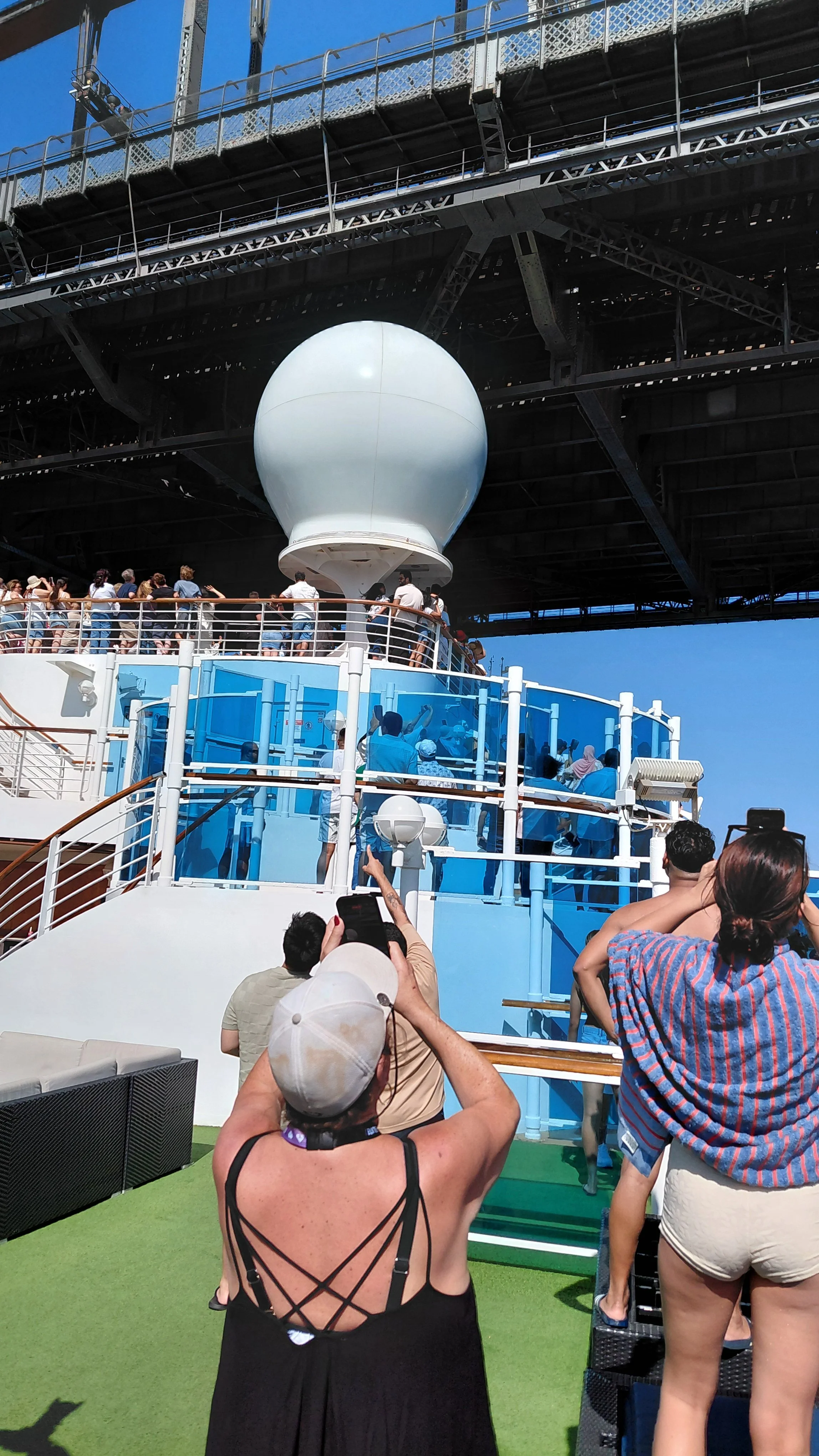 People on the deck of a cruise ship with a large white sphere structure underneath an overhang, likely an observation or communication device, with blue sky in the background.
