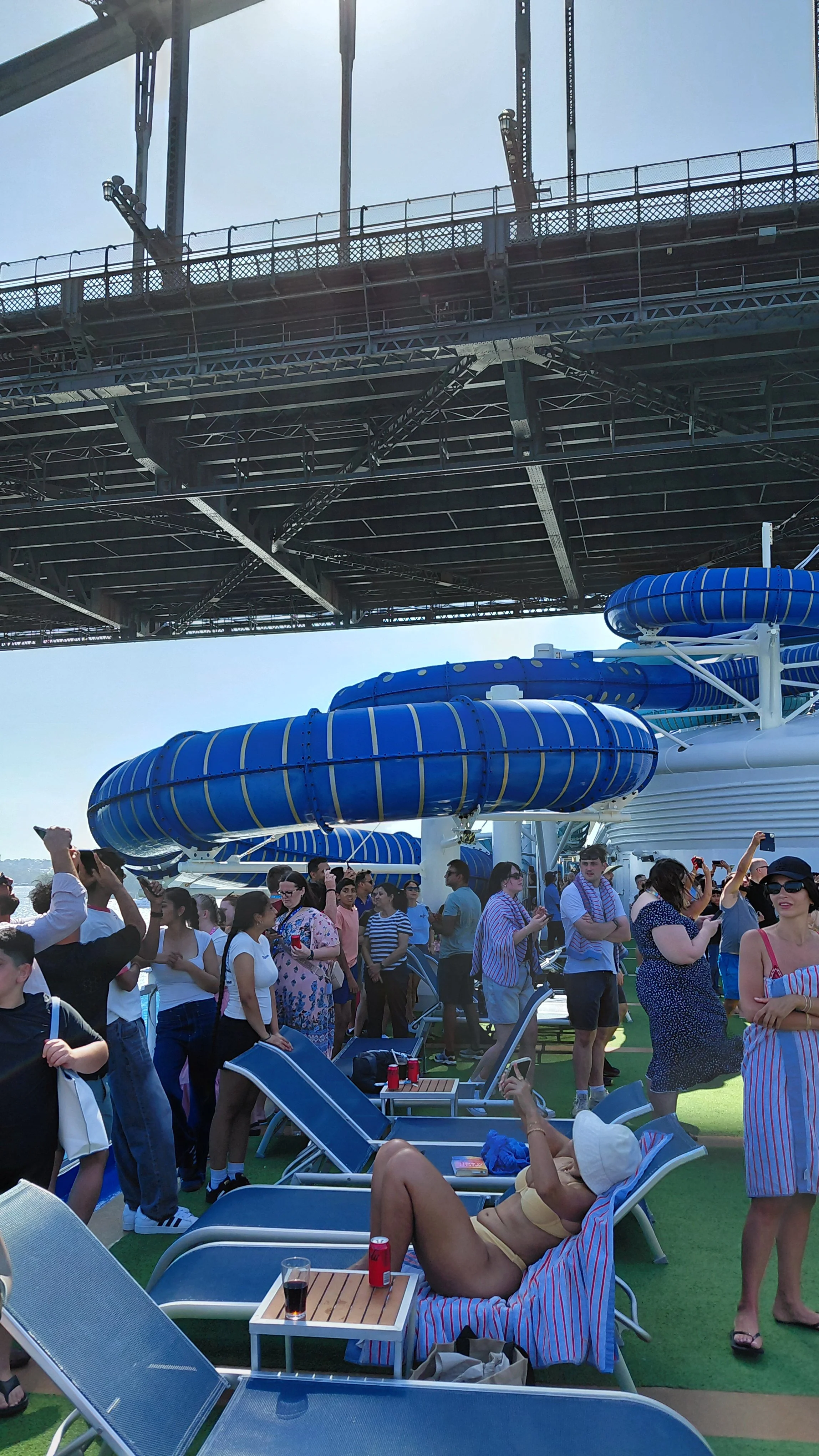 Crowd of people relaxing and taking photos on the deck of a cruise ship with water slides and a large metal structure overhead.