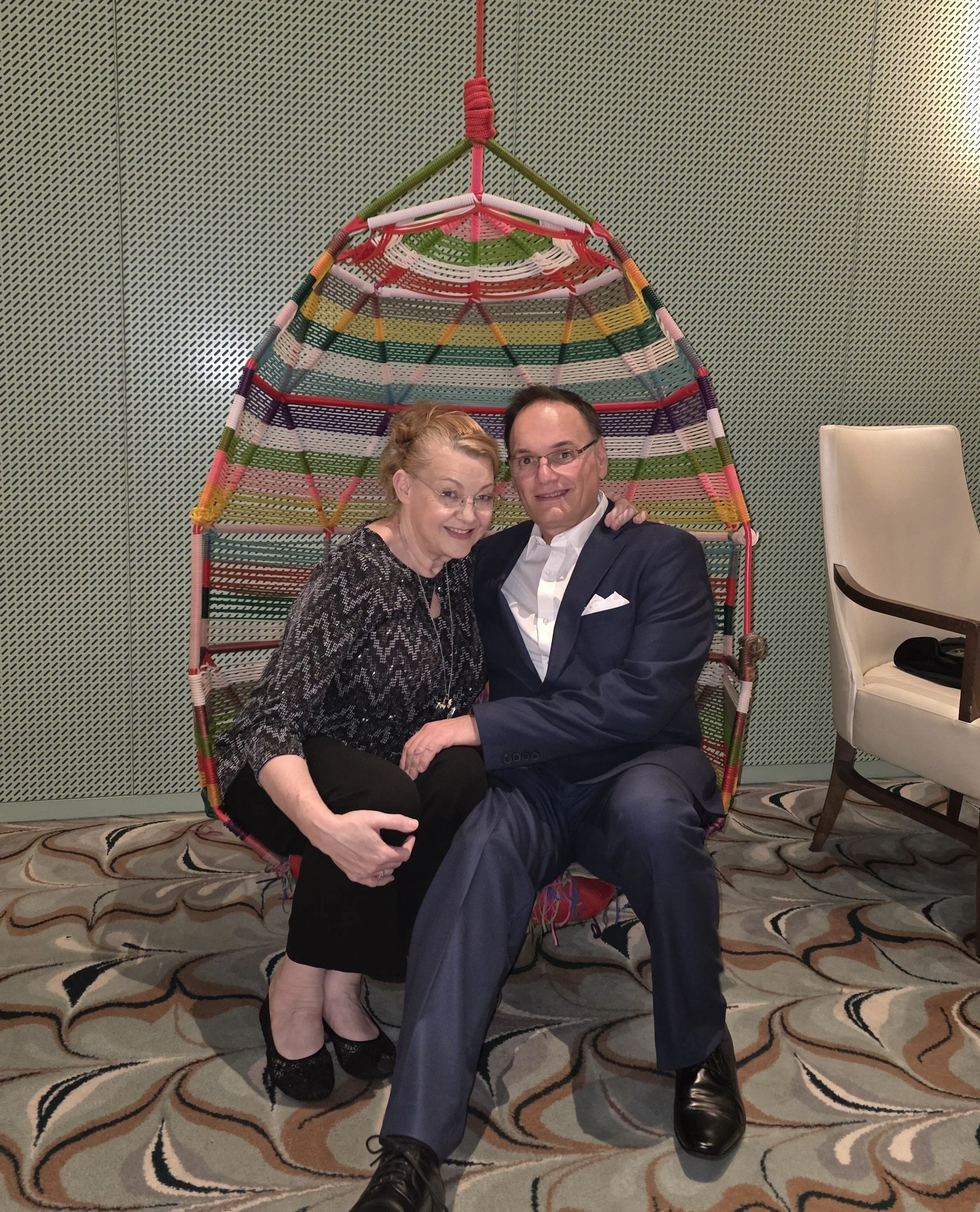 A smiling woman and man sitting together on a colorful woven hanging chair in a room, with one arm around each other.