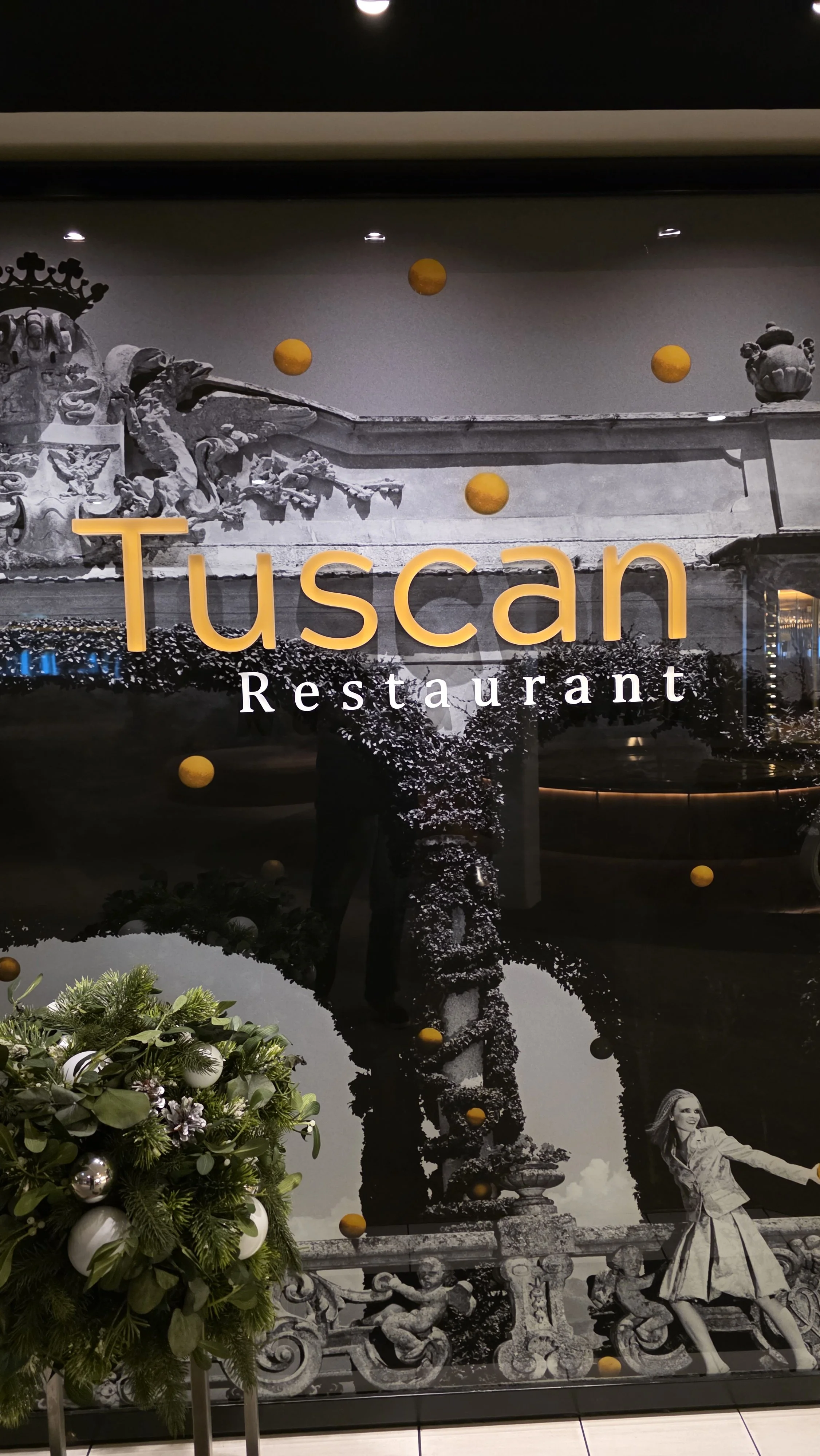 A window sign for Tuscan Restaurant featuring black-and-white images of classical sculptures and architecture, with the restaurant name in orange and white lettering, and a small floral arrangement in front.