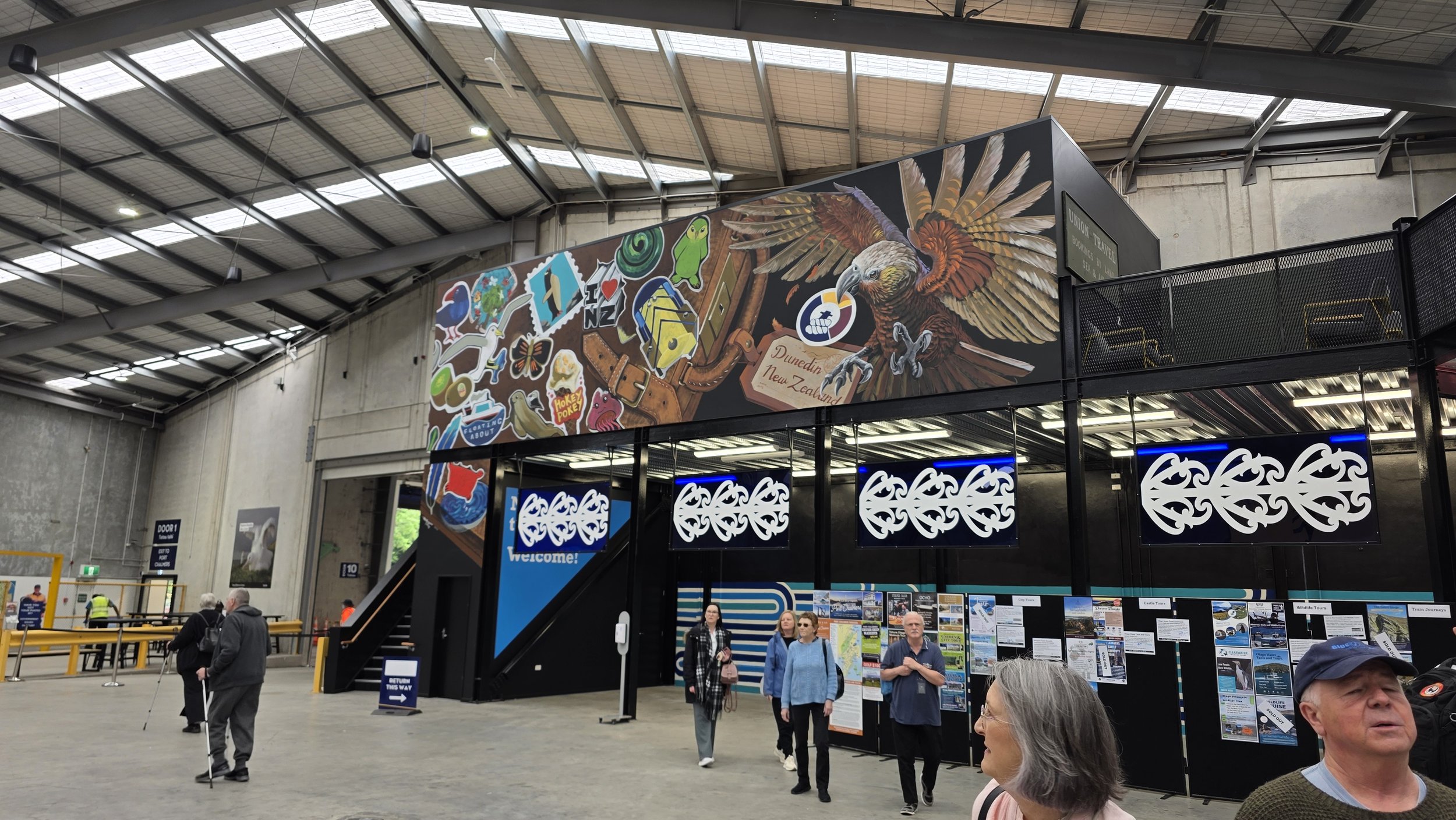 Interior of a train station with people walking and signs. A large colorful mural promotes Dunedin, New Zealand, featuring a bird, kites, and various stickers and graphics.