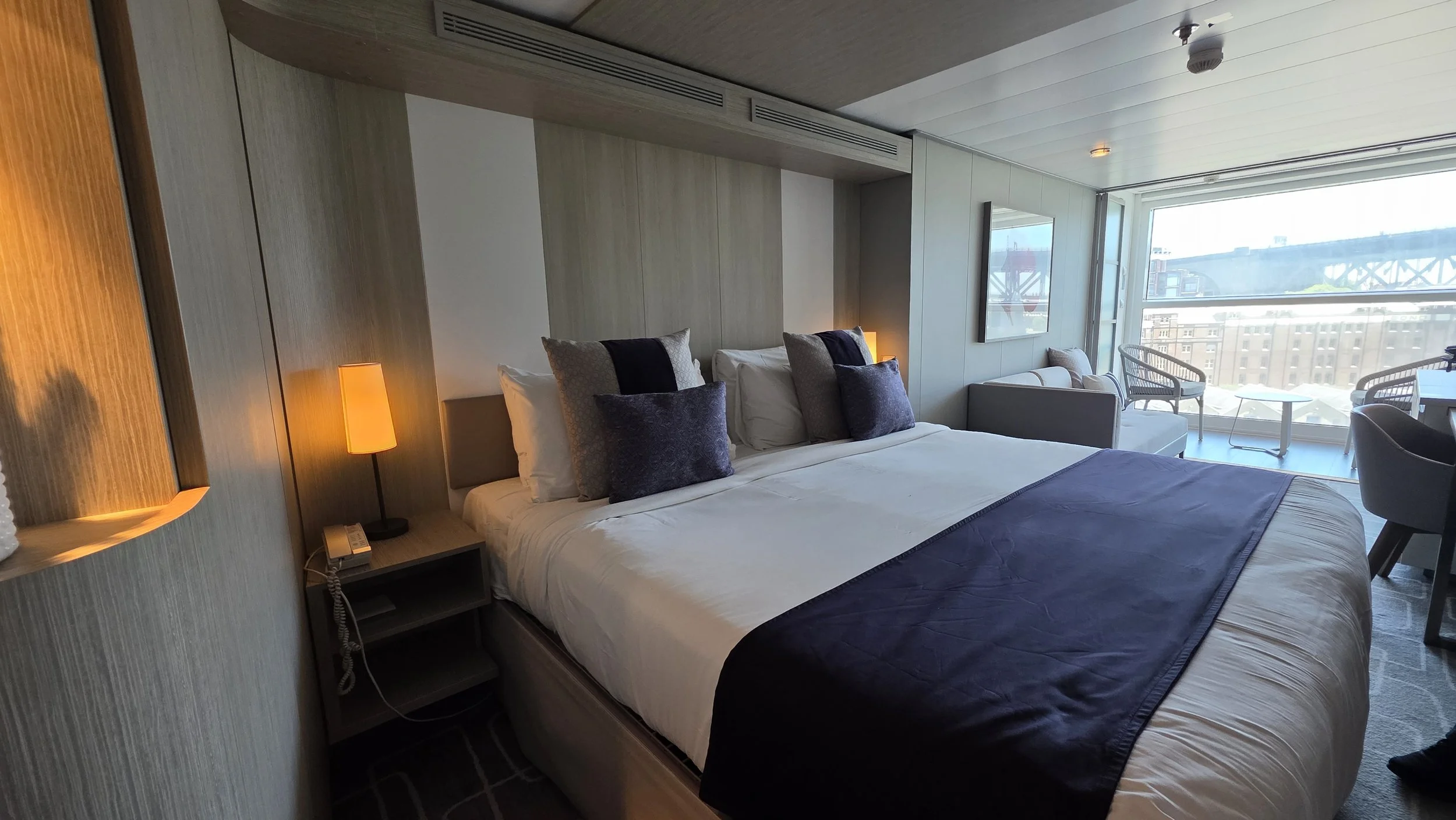 A hotel room with a large bed, decorative pillows, nightstand with a lamp and telephone, and a sitting area with chairs and a table near the window showing city buildings and a bridge.