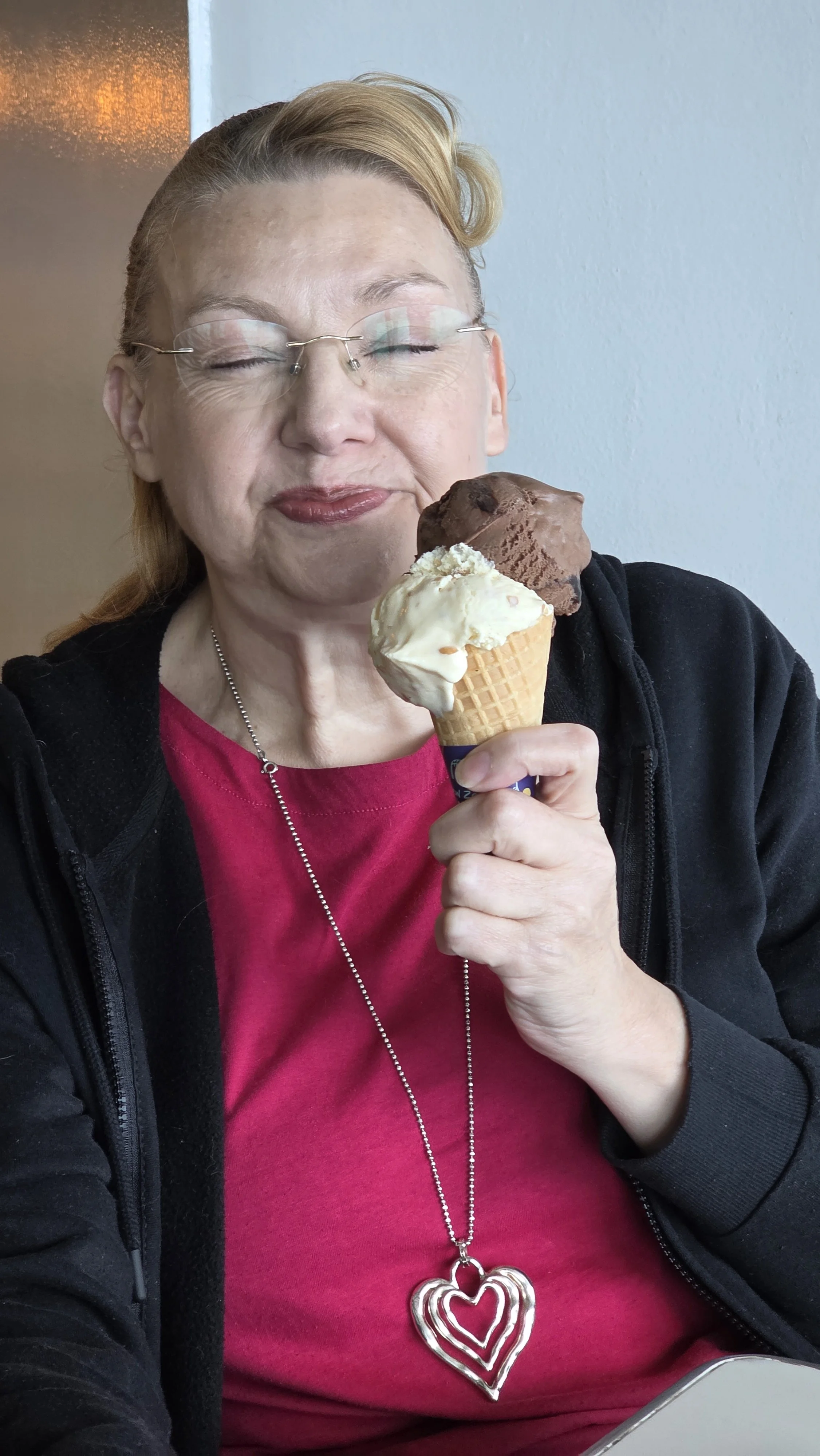 A woman with glasses, red hair, and a red shirt holding two scoops of ice cream in a cone, one chocolate and one vanilla, smiling with her eyes closed.