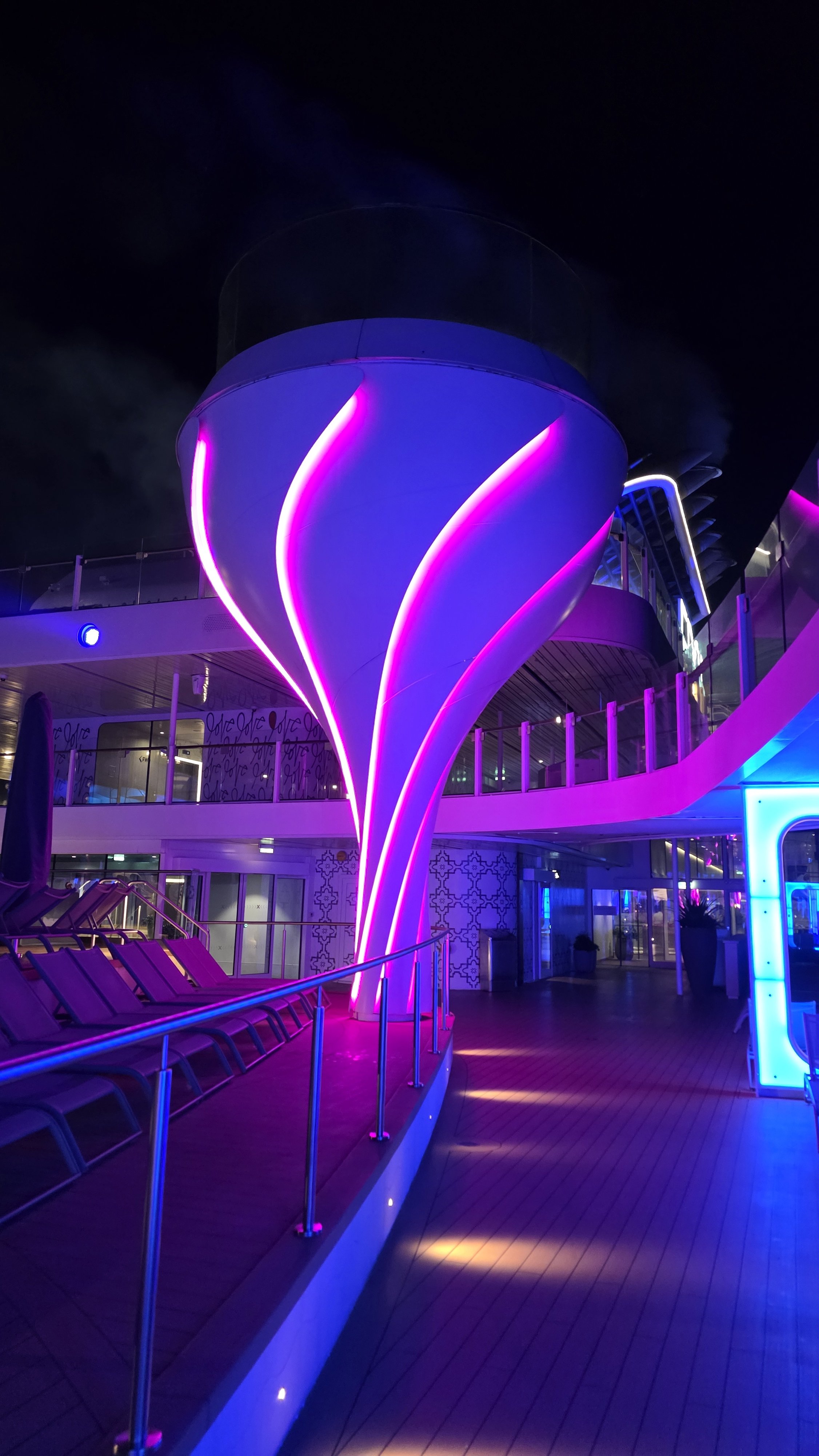Night view of a cruise ship deck with vibrant purple and pink illuminated striped light sculpture resembling an abstract tree or plant, with lounge chairs and glass railings.