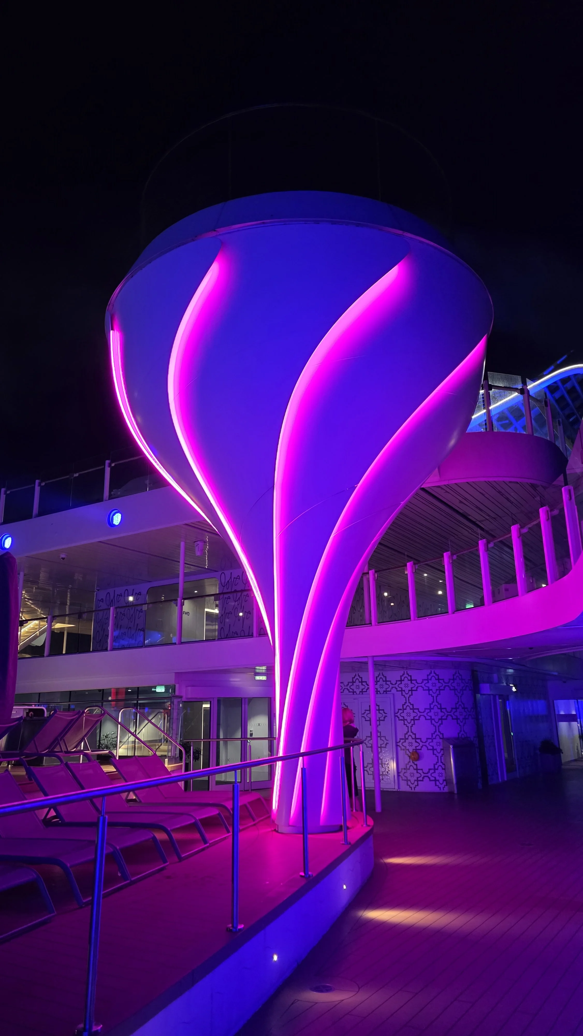 Nighttime scene on a cruise ship or waterfront with illuminated purple and pink sculptural structures, lounge chairs, and a balcony overlooking the water.