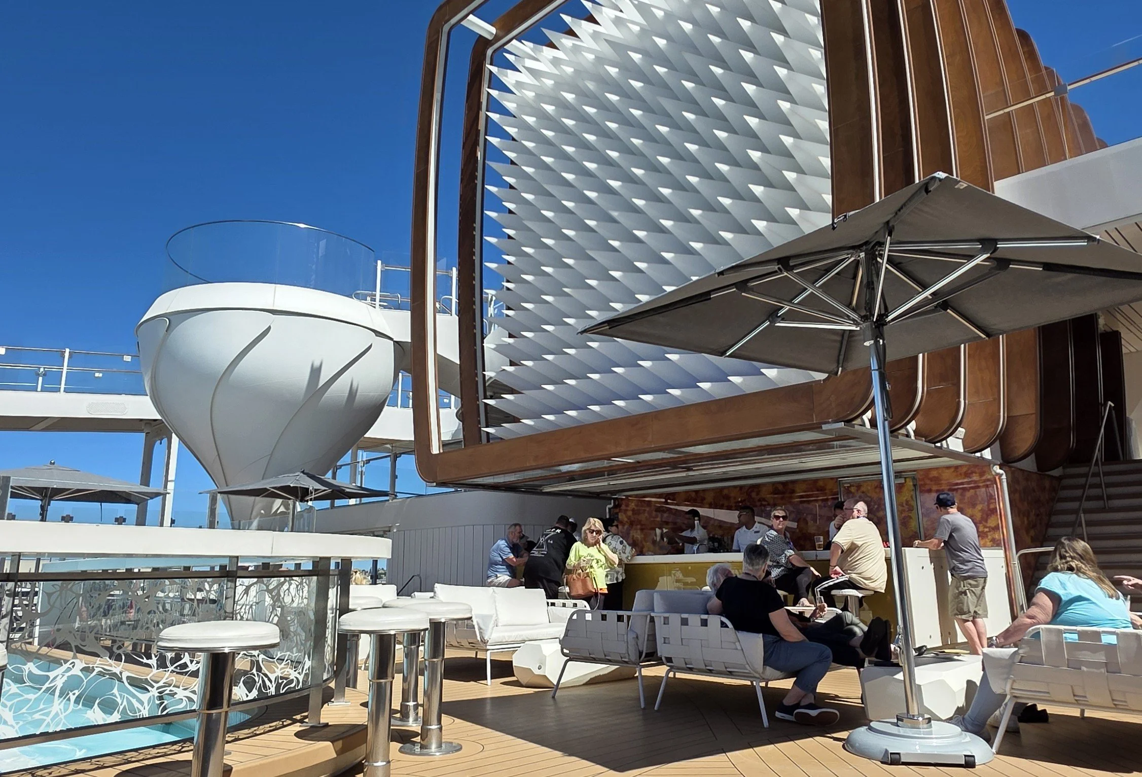 People relaxing and socializing on the deck of a cruise ship under large umbrellas, with modern architectural features and a clear blue sky in the background.