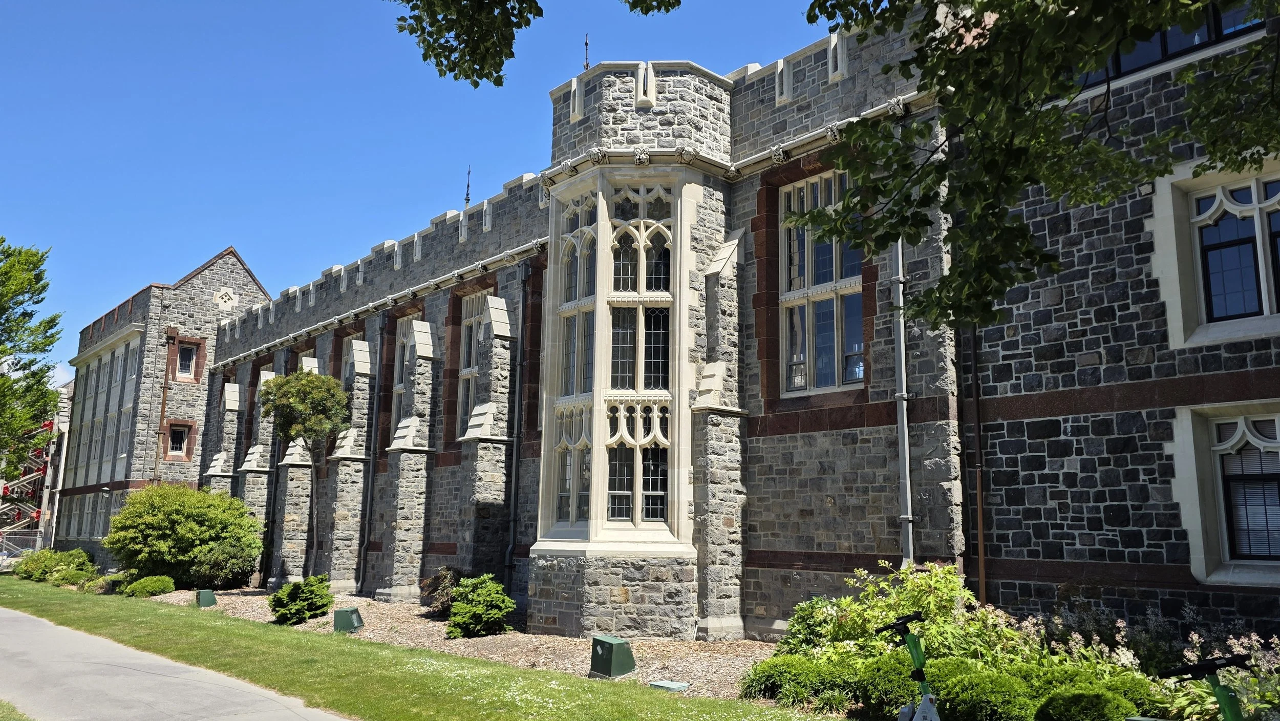 A historic stone building with gothic-style windows, surrounded by greenery and a sidewalk, under a clear blue sky.
