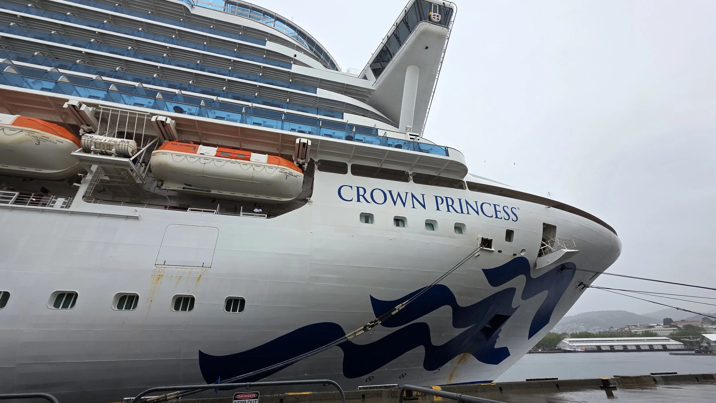 Close-up of the bow of the cruise ship Crown Princess docked at a port, showing safety lifeboats and the ship's name.