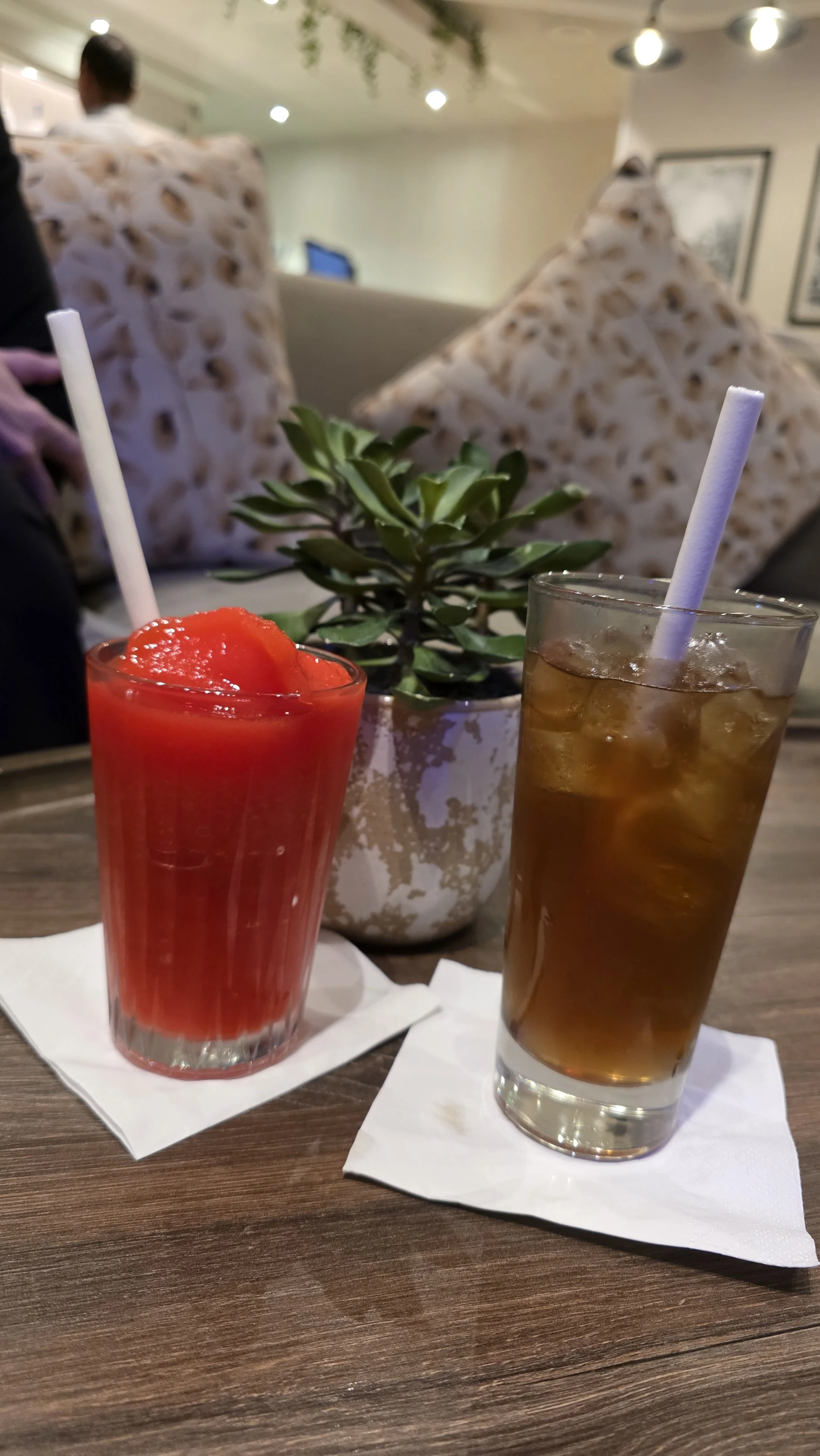 Two drinks on a wooden table, one red fruit smoothie and one iced tea, with a potted green plant behind them, in a café or restaurant setting.