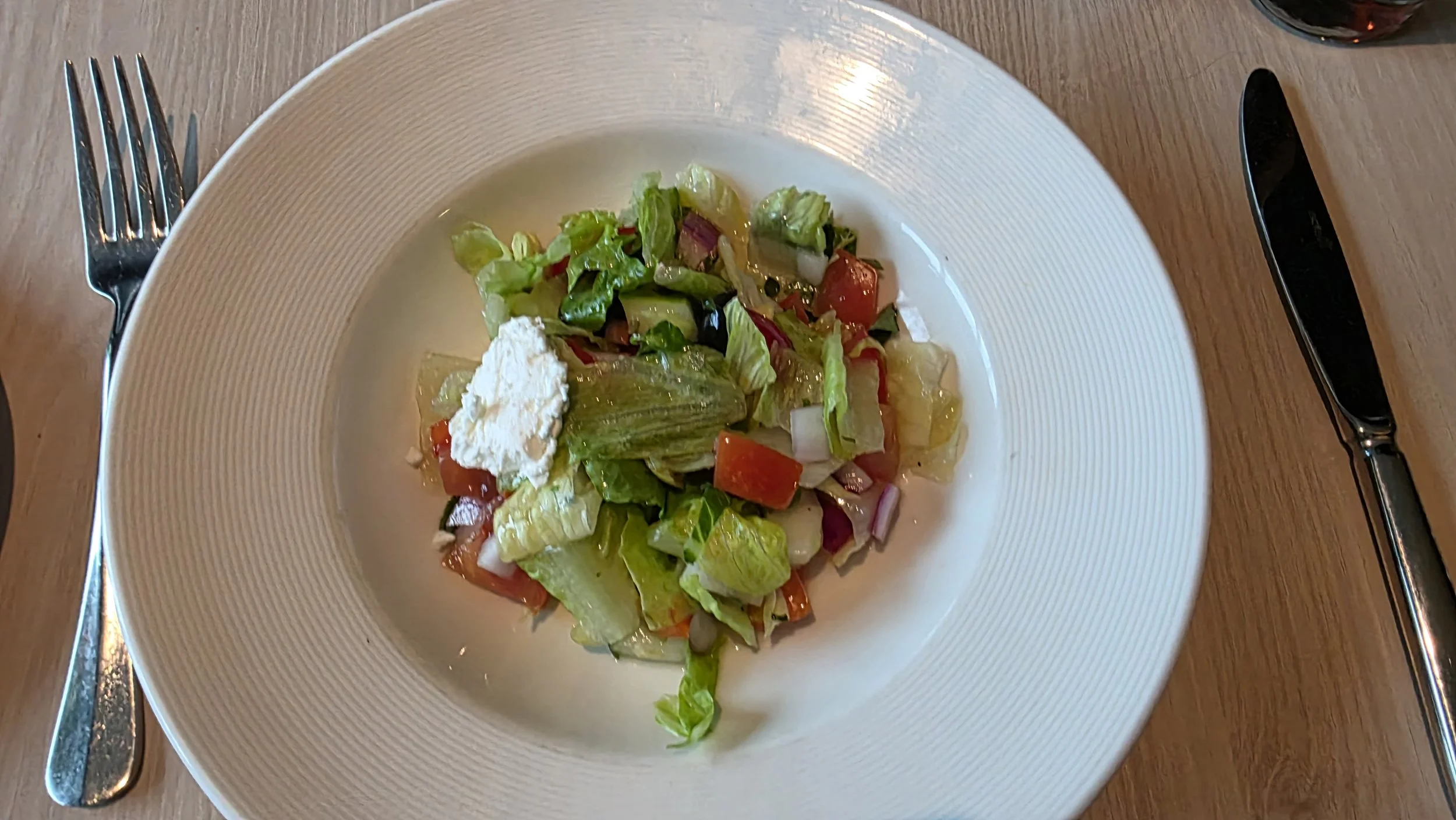 A plate of mixed salad with lettuce, tomato, onion, and cottage cheese, served with a fork on the left and a knife on the right on a wooden table.