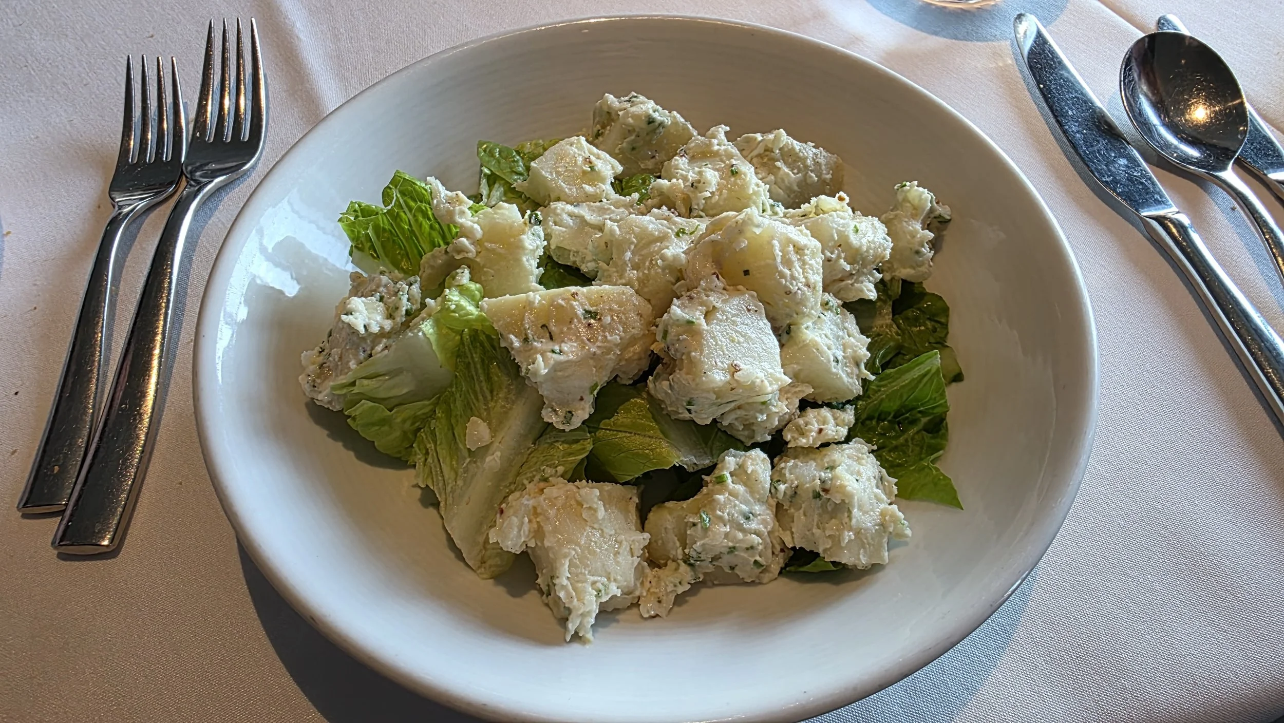 A bowl of potato and egg salad on a table with silverware on either side.