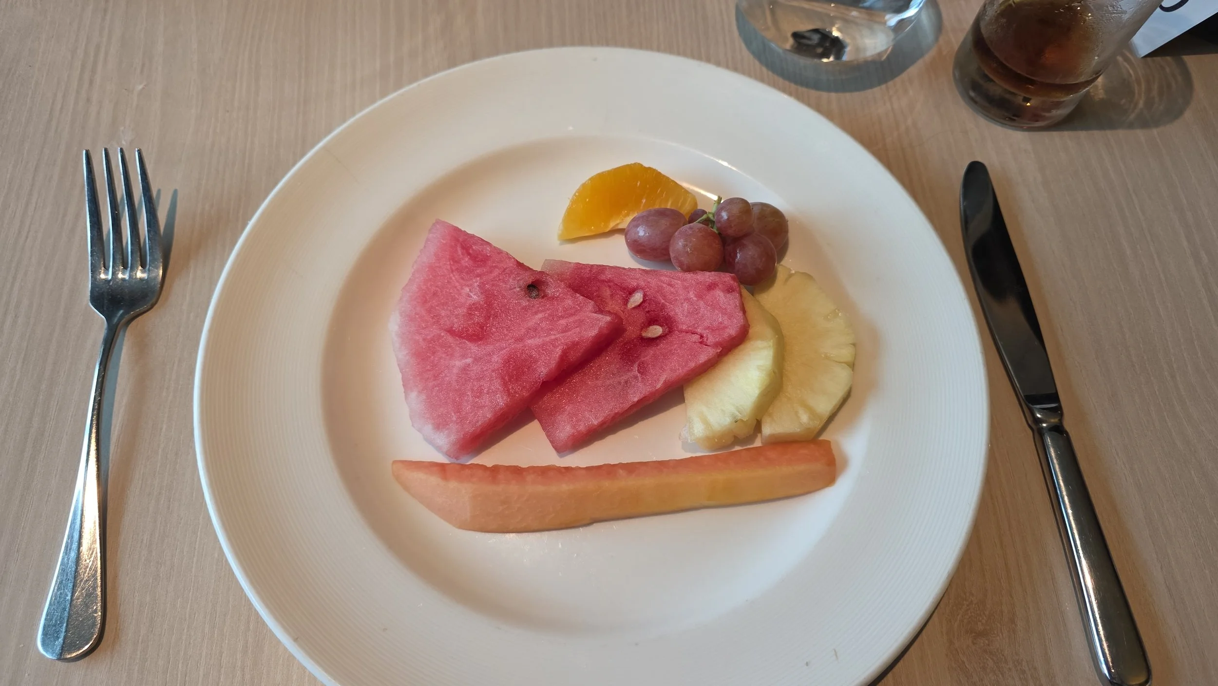 A white plate with slices of watermelon, pineapple, cantaloupe, apple, a cluster of grapes, and a wedge of orange on a wooden table, with a fork on the left and a knife on the right.