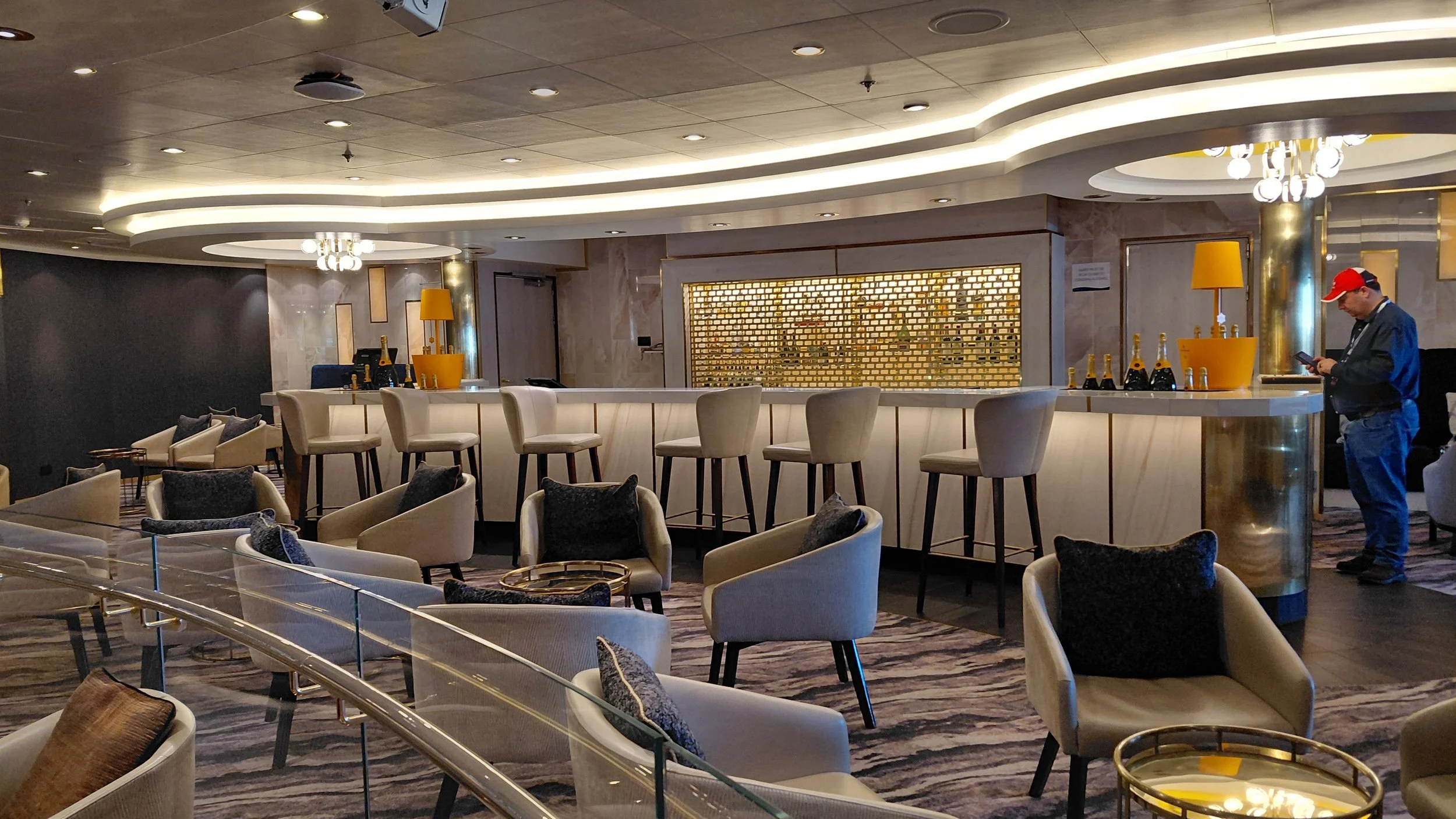 An upscale hotel bar with a curved white bar counter, beige chairs, and tables with black cushions. There is a man in a red cap using his phone, and bottles of champagne on the counter.