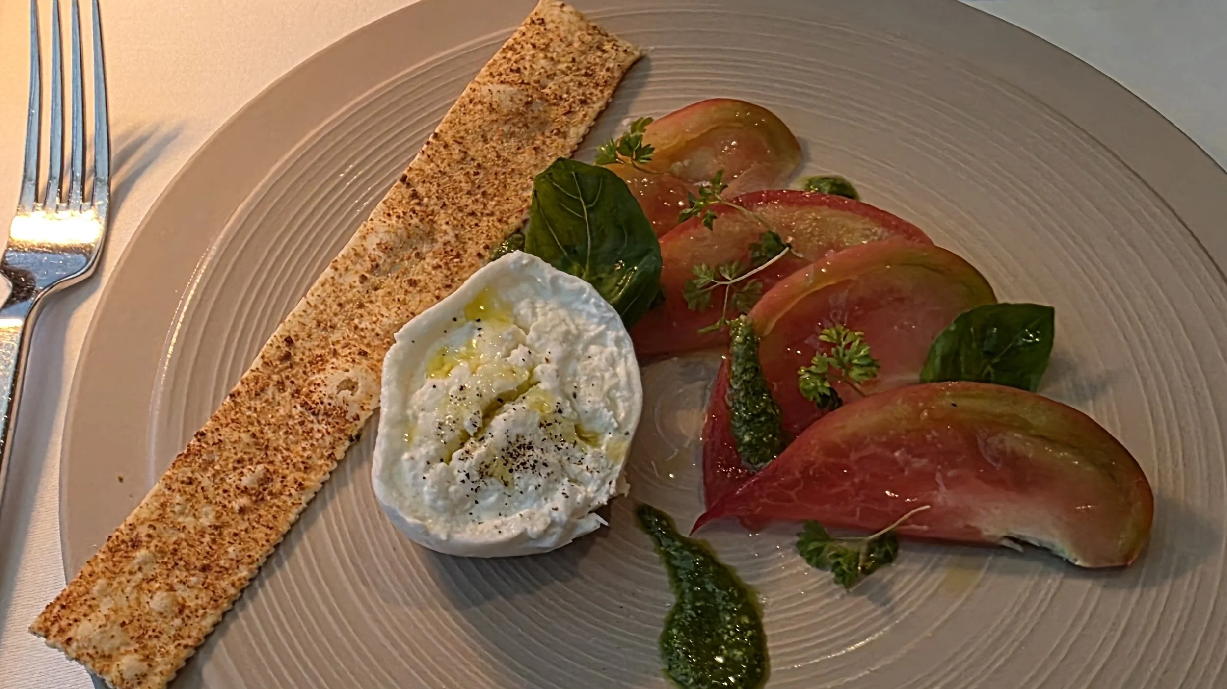 A plate of sliced heirloom tomatoes garnished with fresh herbs, served with a scoop of burrata cheese, a green sauce, and a long crispy breadstick.