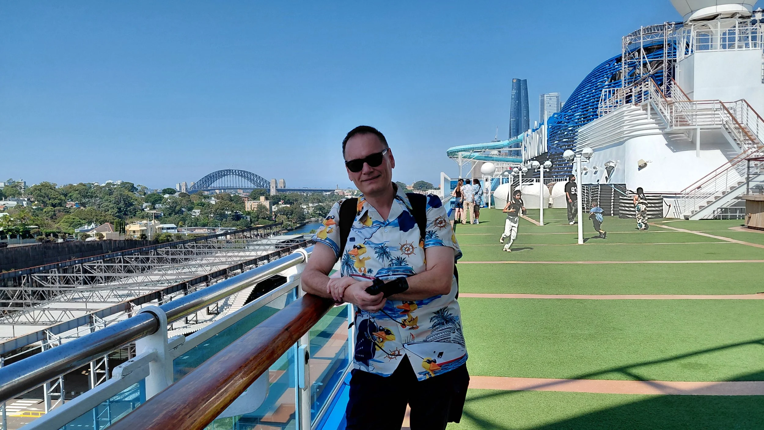 A man wearing a colorful tropical shirt and sunglasses leaning on a rail on the deck of a cruise ship, with a city skyline, a bridge, and some people in the background on the ship's top deck under a clear blue sky.