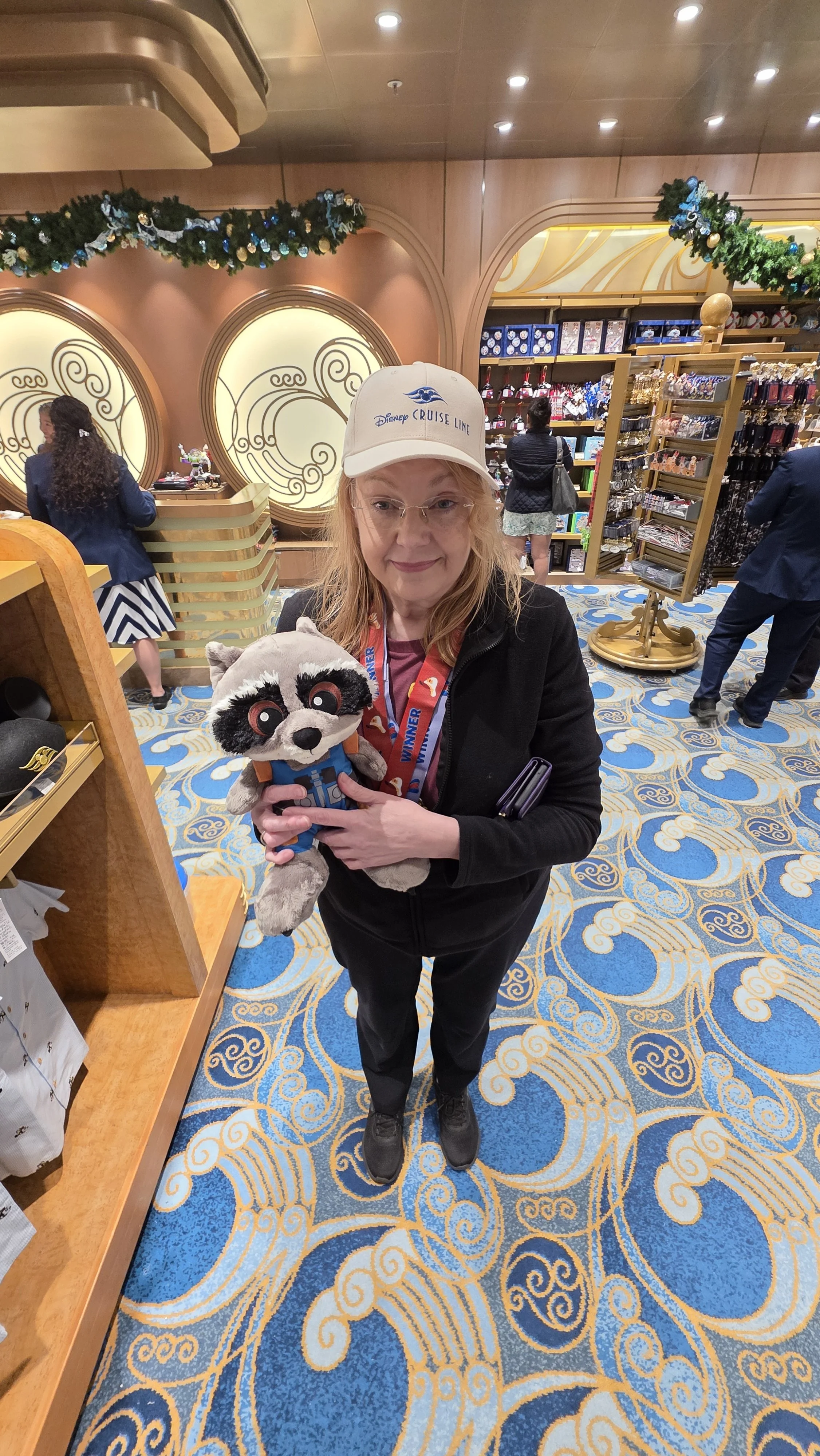 A woman wearing a Disney Cruise Line cap standing in a gift shop, holding a Rocket Raccoon plush toy from Guardians of the Galaxy, with shelves of merchandise in the background.