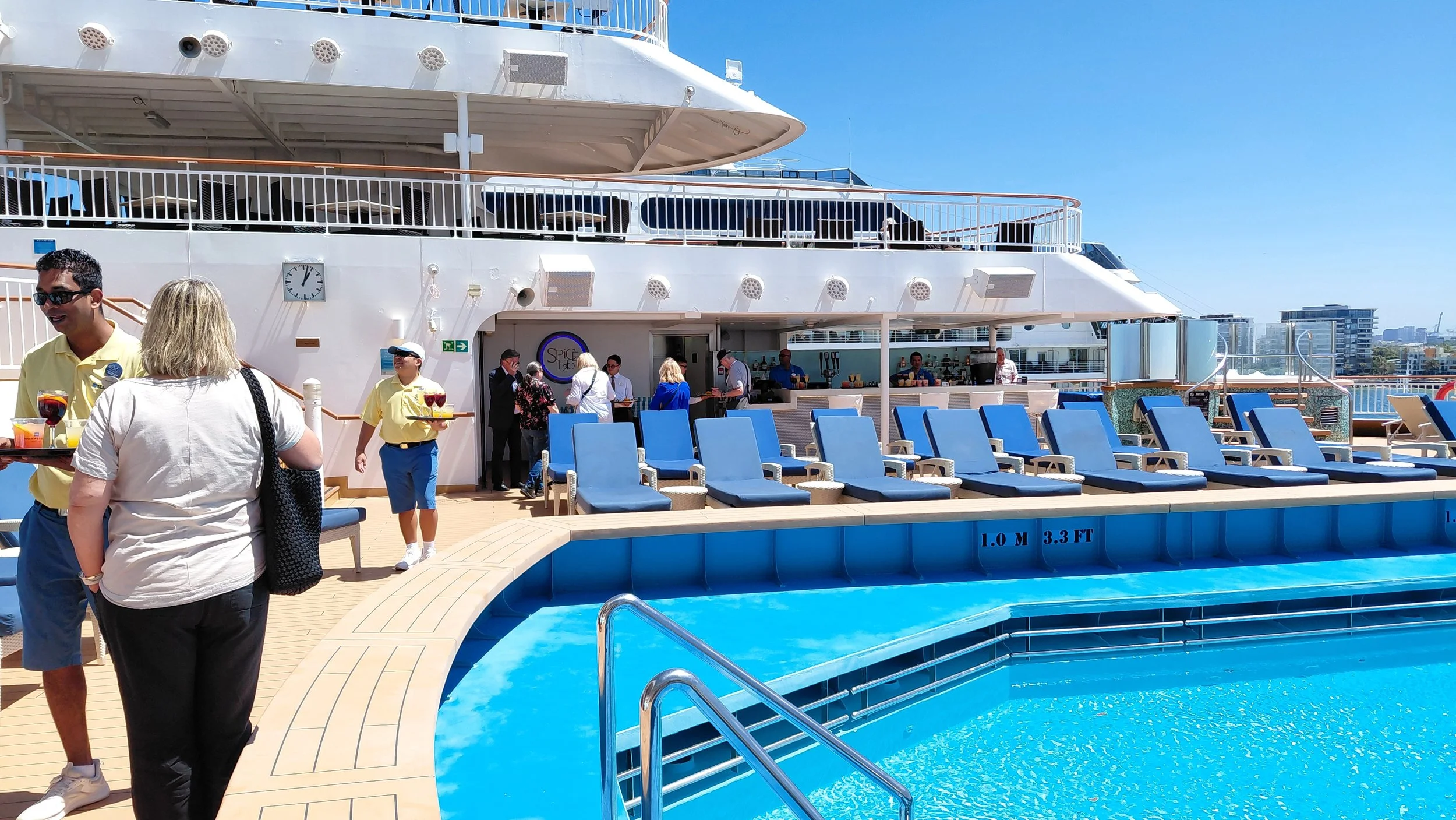 People on a cruise ship deck near a swimming pool, some service staff assisting passengers, and city buildings in the background on a sunny day.