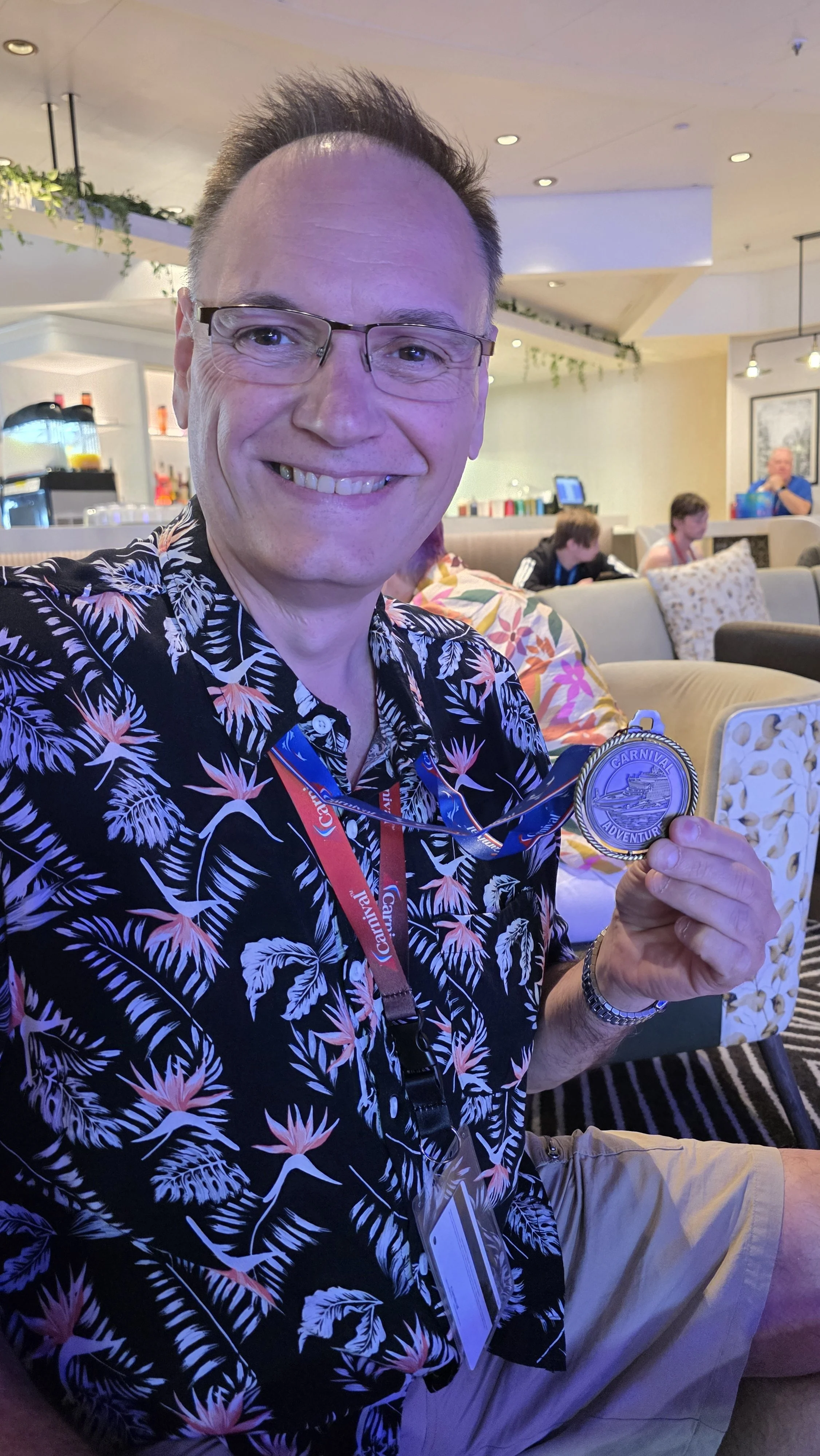 A man smiling while holding a silver medal that says 'Carnival Adventure' in a lounge area with patterned chairs and other people in the background.