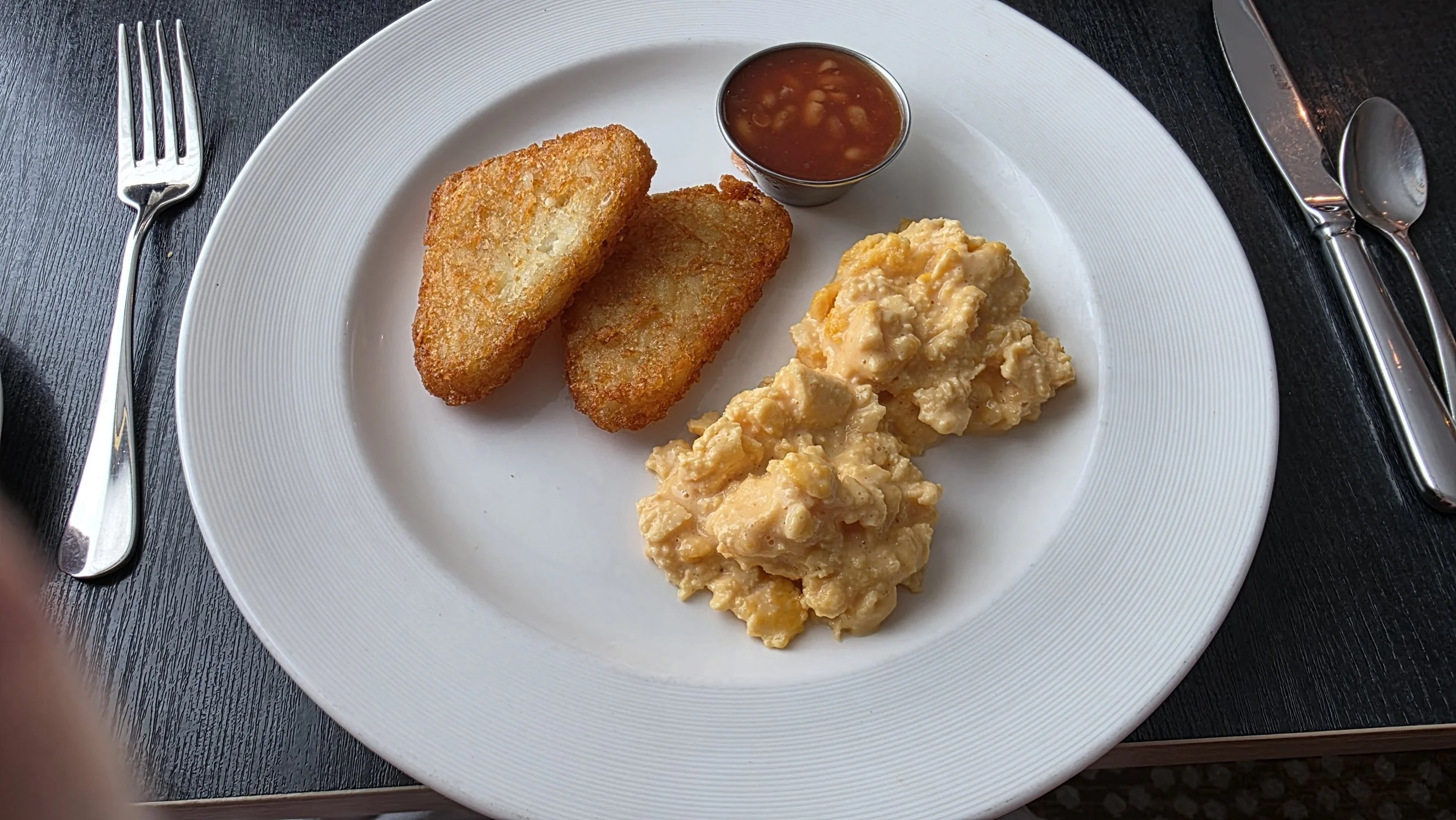 Plate of breakfast with two fried potato slices, scrambled eggs, and a small cup of salsa or sauce.