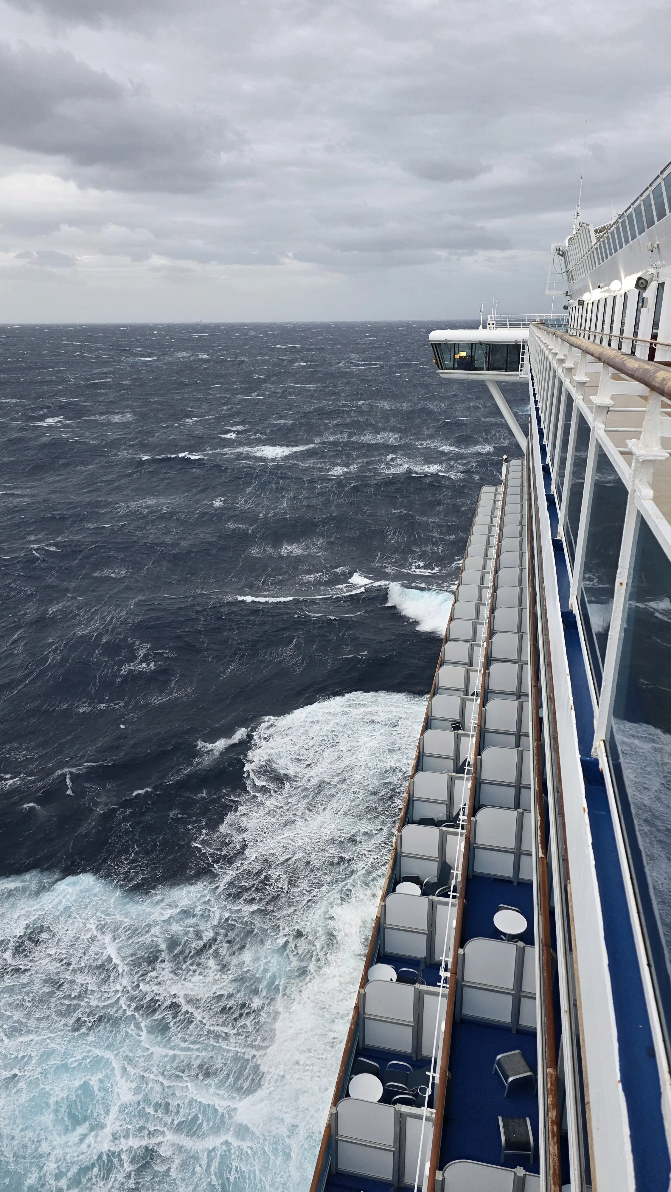 View from a cruise ship looking out at the open ocean with waves and an overcast sky, featuring part of the ship's deck with seats and an observation area.