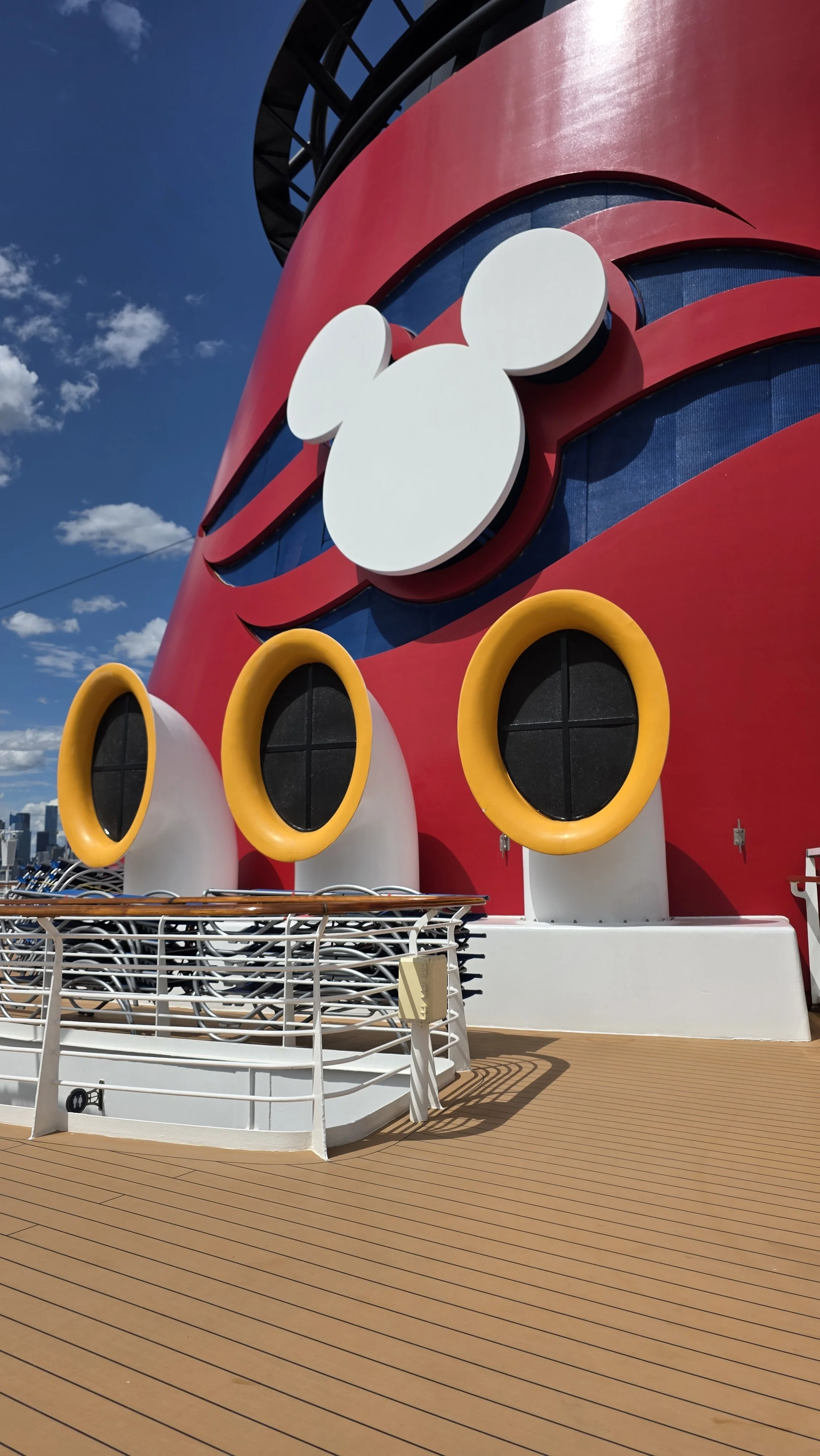 Close-up of the upper part of the Disney Cruise Line ship, featuring the iconic Mickey Mouse head logo in white and portholes with bright yellow rings on a red and blue background.