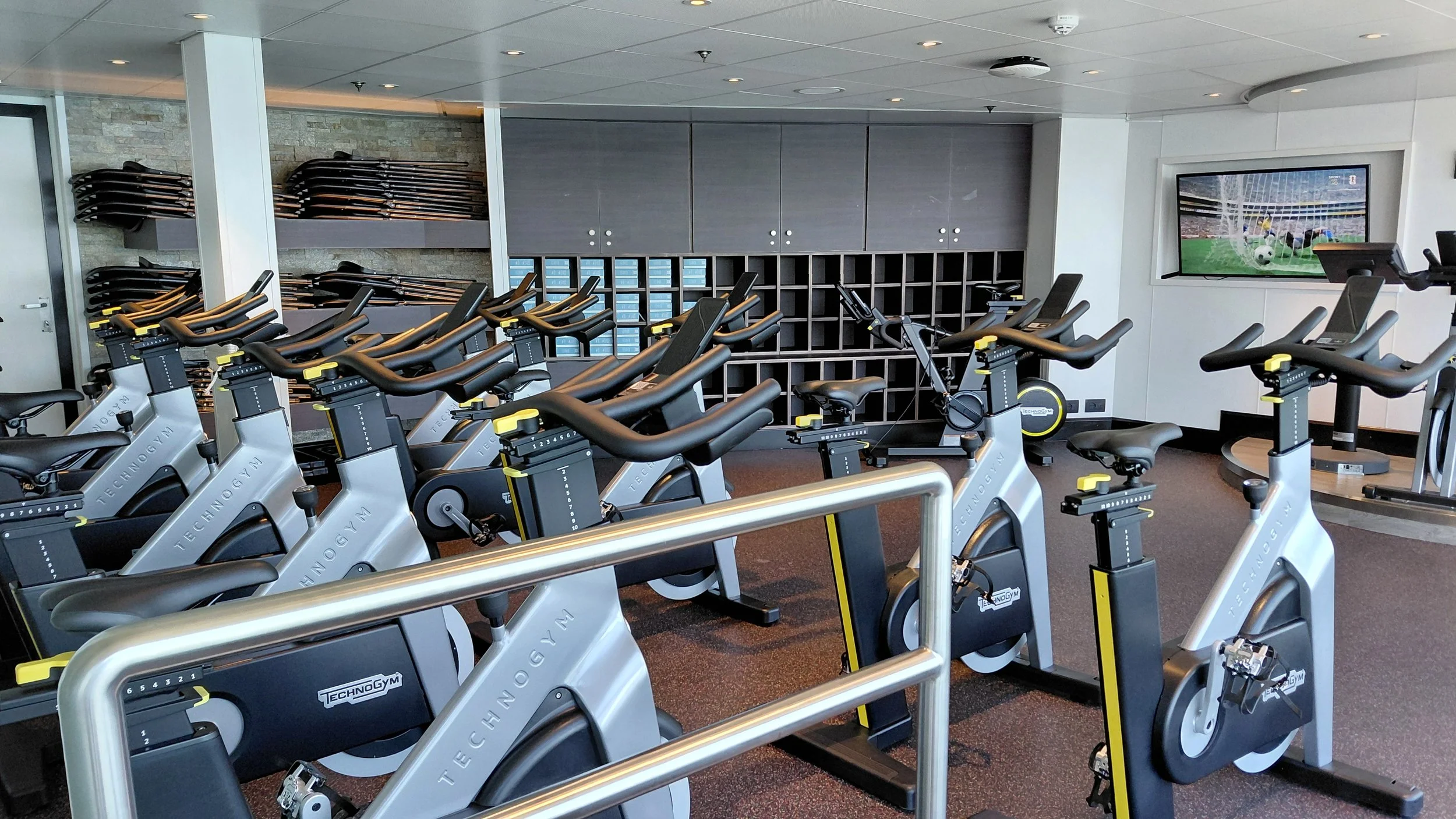 Indoor gym with multiple stationary bikes arranged in rows, a television screen on the wall showing a soccer game, and stacked exercise mats on shelves in the background.