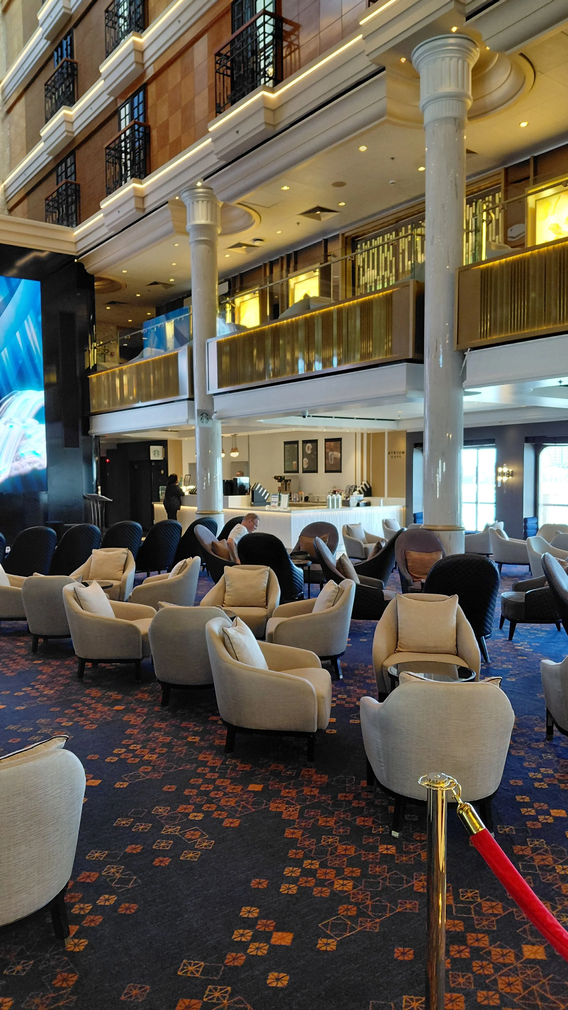 An elegant hotel lobby with numerous beige and dark-colored armchairs, some with pillows, arranged for seating. The space features large columns, a patterned carpet with orange and dark hues, and a reception counter in the background. There are a few