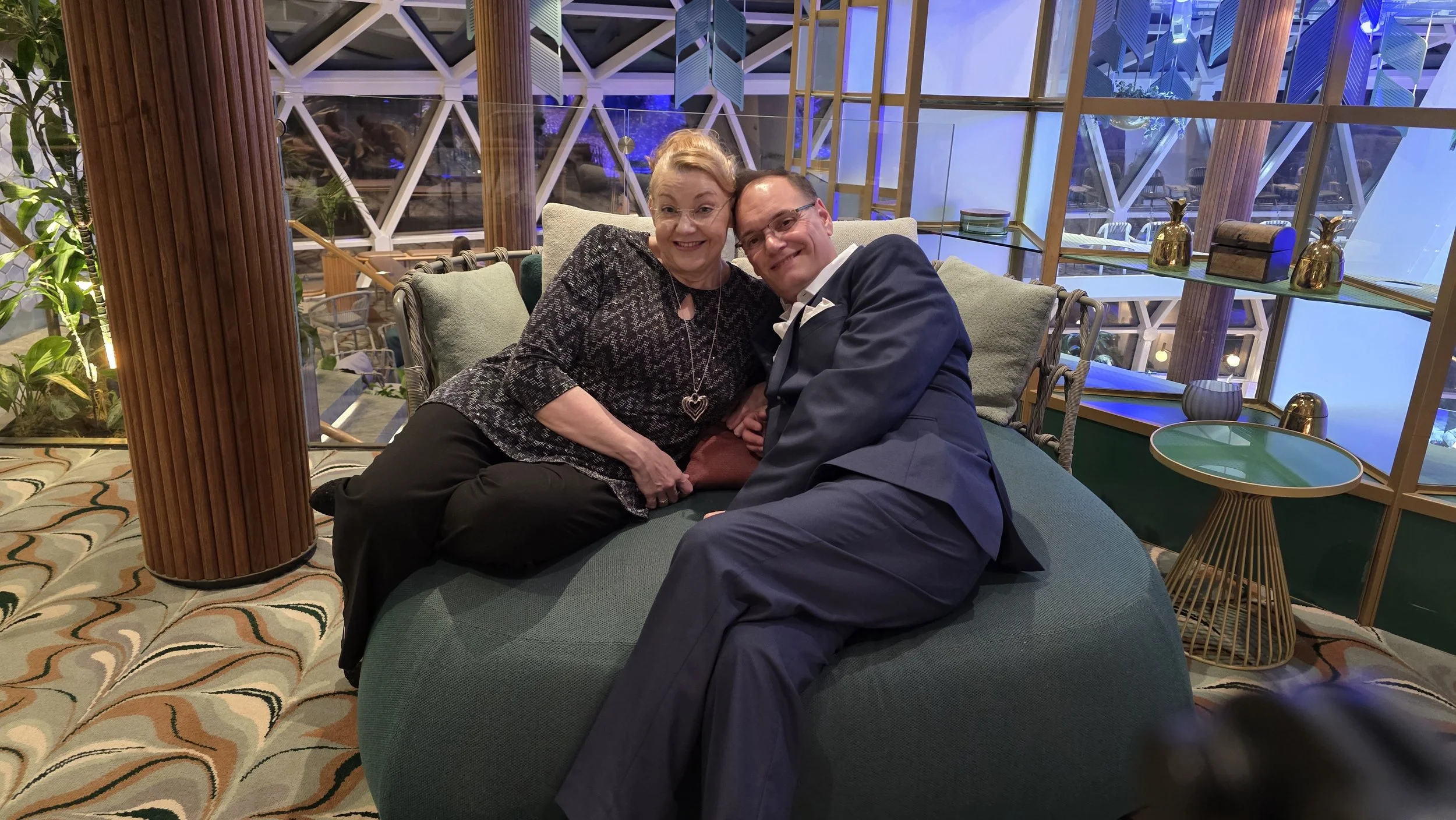 An elderly woman and a middle-aged man sit closely together on a lounge chair, smiling warmly at the camera. The woman wears a black patterned top and a heart-shaped necklace, while the man is dressed in a dark suit with a white shirt. They appear ha